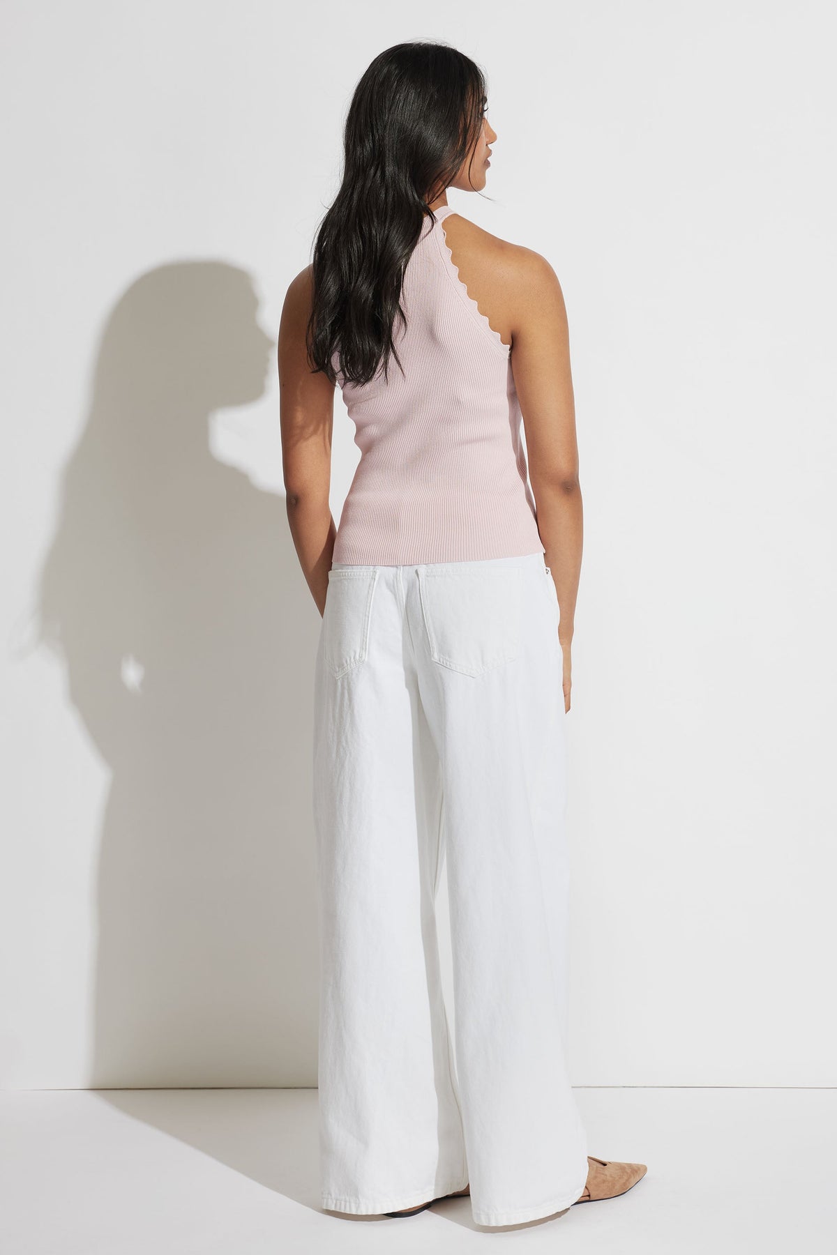 Woman wearing a soft pink scalloped halter tank paired with wide-leg white pants, shown from behind against a white wall.