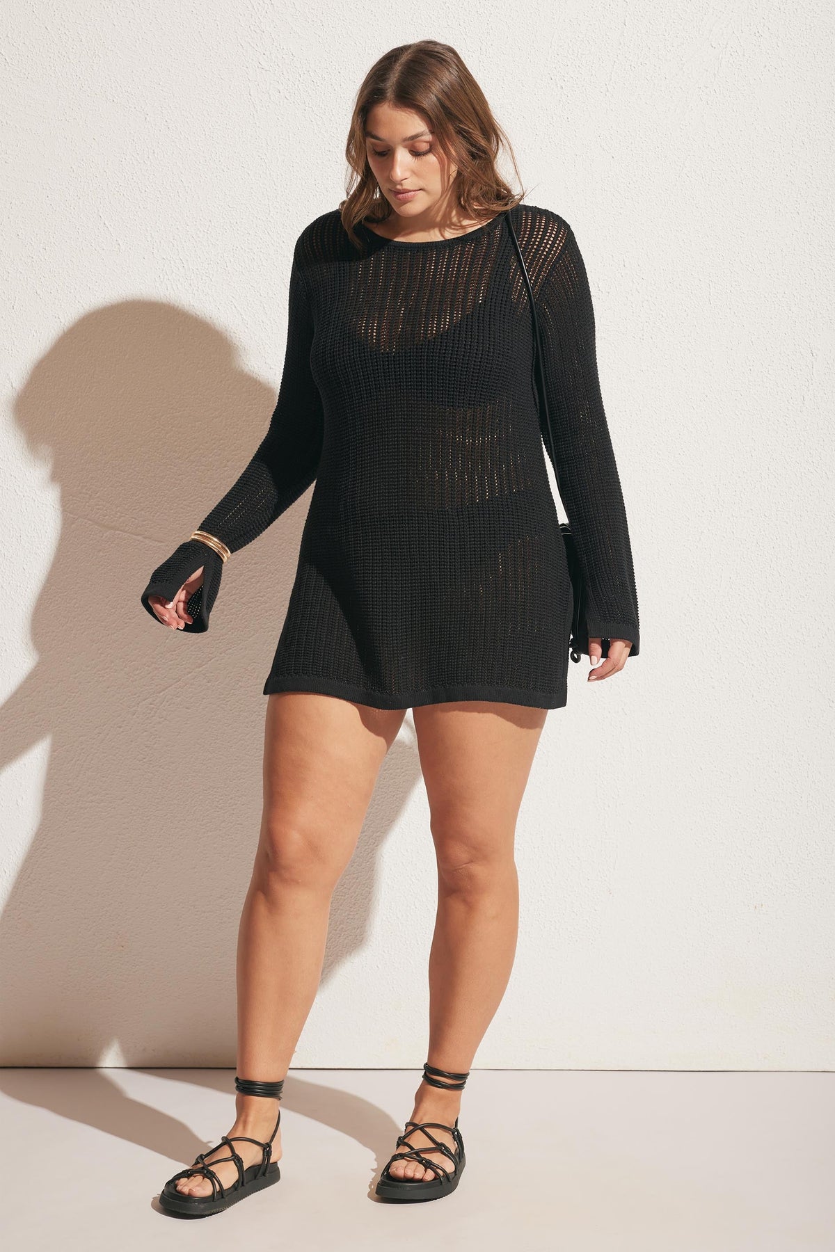 Woman wearing a black crochet tunic dress with long sleeves and lace-up sandals against a plain wall