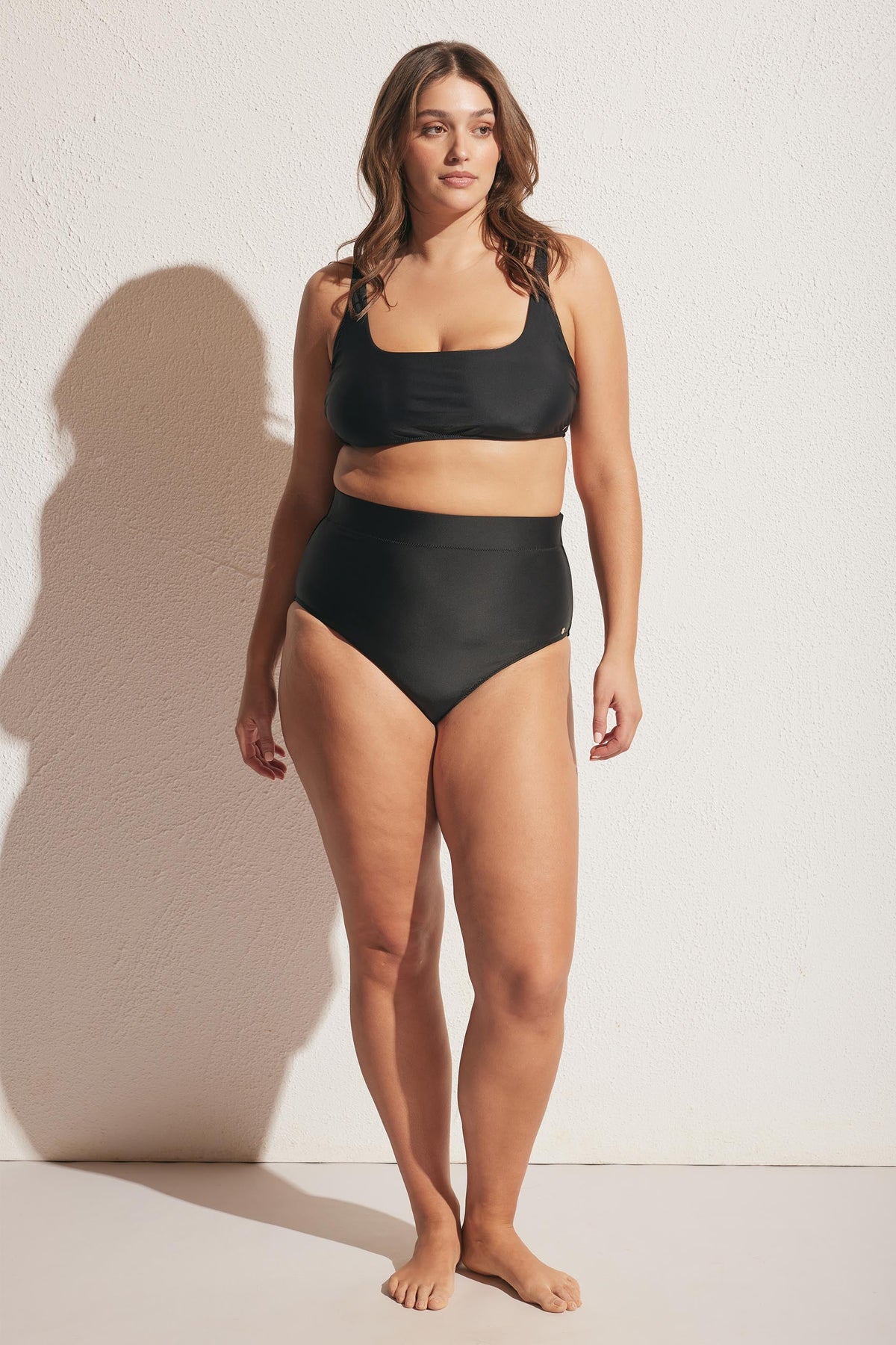 Woman standing in a matte black square neck bikini top with matching high-waisted bikini bottoms against a white wall
