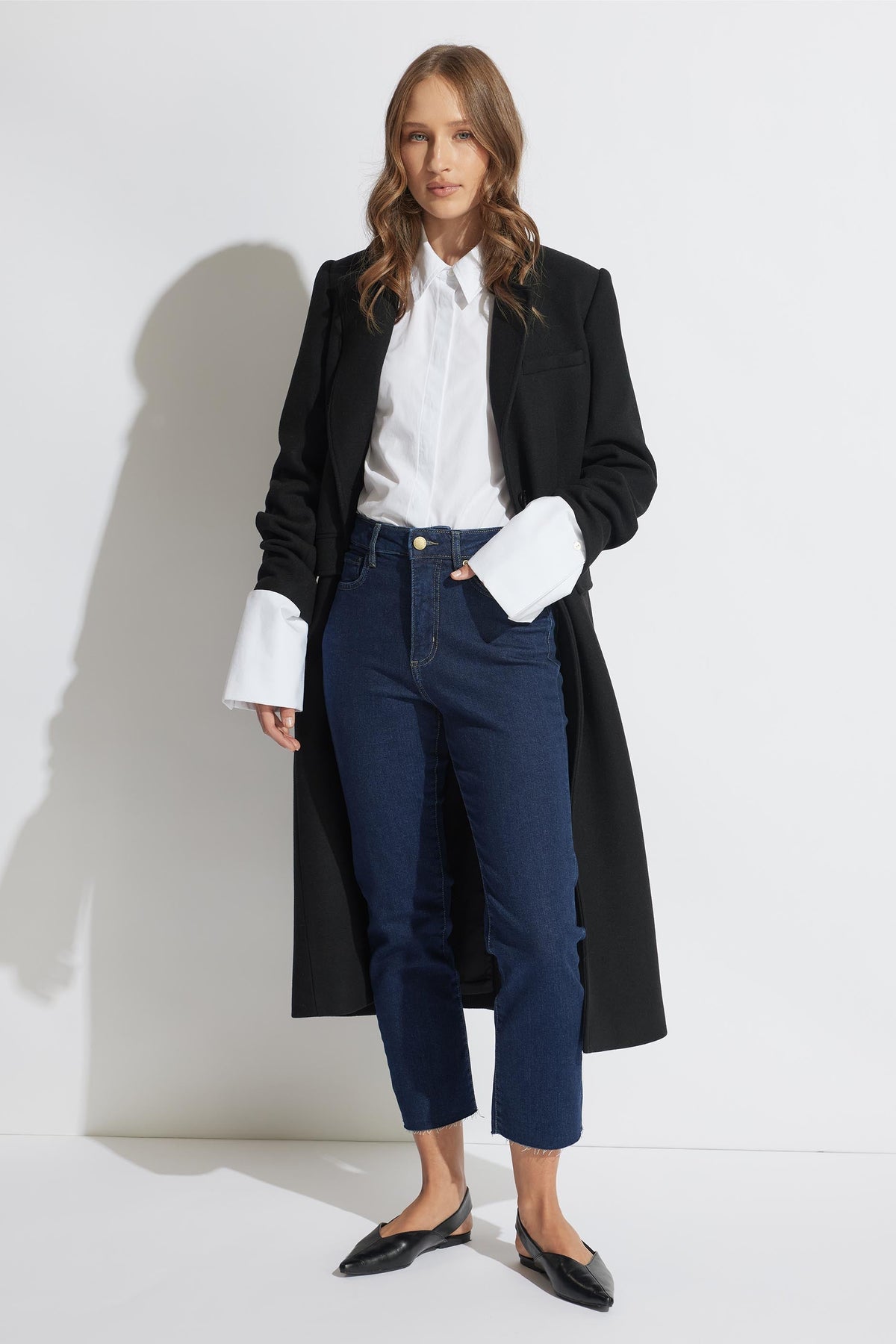 A model is wearing a high-rise, straight-leg crop jeans in a dark wash denim. She has a white shirt with long sleeves, black coat, and carrying a black bag with a chain shoulder strap.