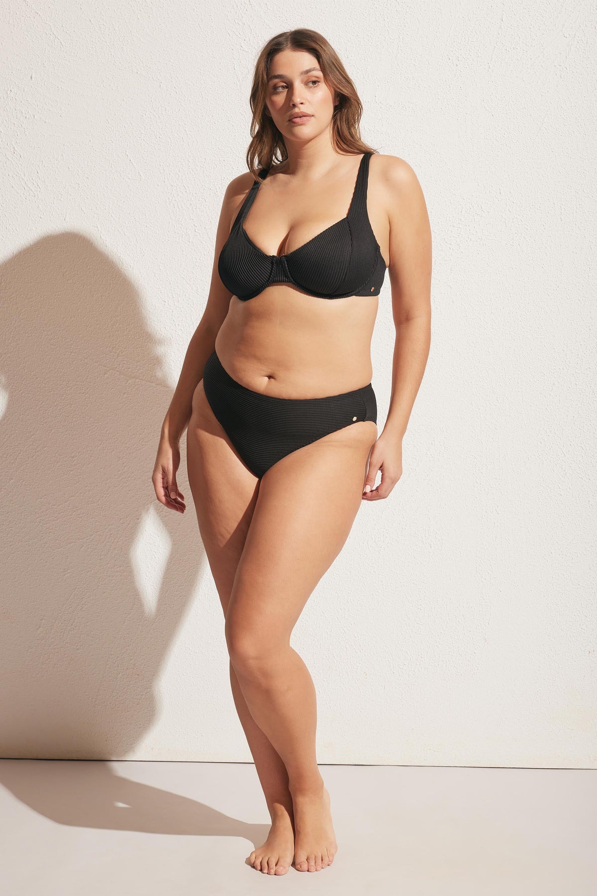 Woman wearing a black ribbed bikini bottom with a supportive underwire top, standing against a textured white wall in natural light