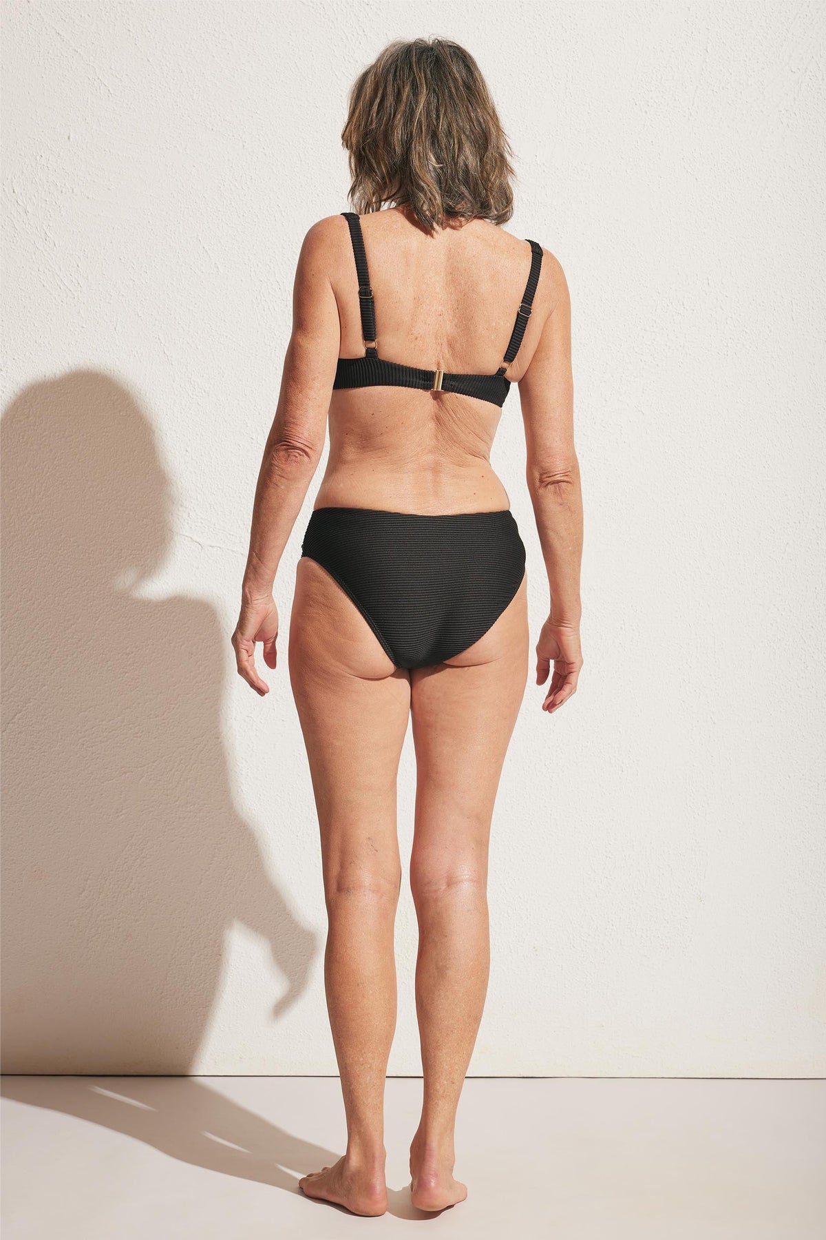 Woman wearing a classic black ribbed bikini bottom with adjustable straps on the matching top, shown from the back.