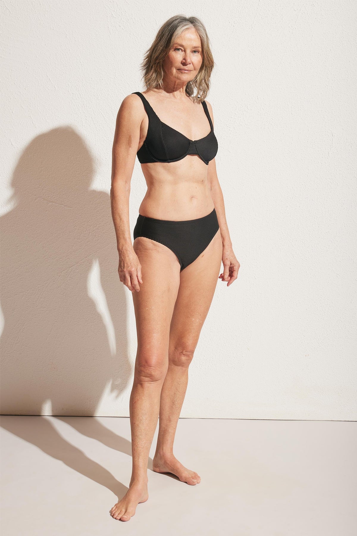 Mature woman wearing a sleek black ribbed bikini bottom and matching underwire bra, standing in natural light