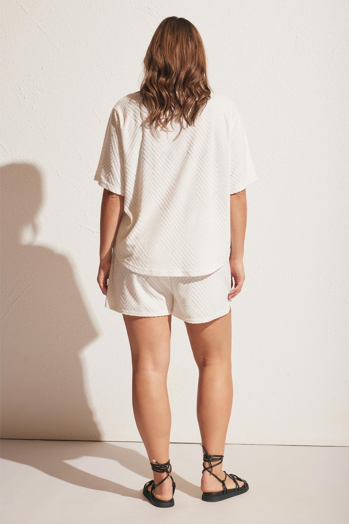 Woman wearing a relaxed-fit white terry fabric shirt and matching shorts paired with black lace-up sandals, seen from behind