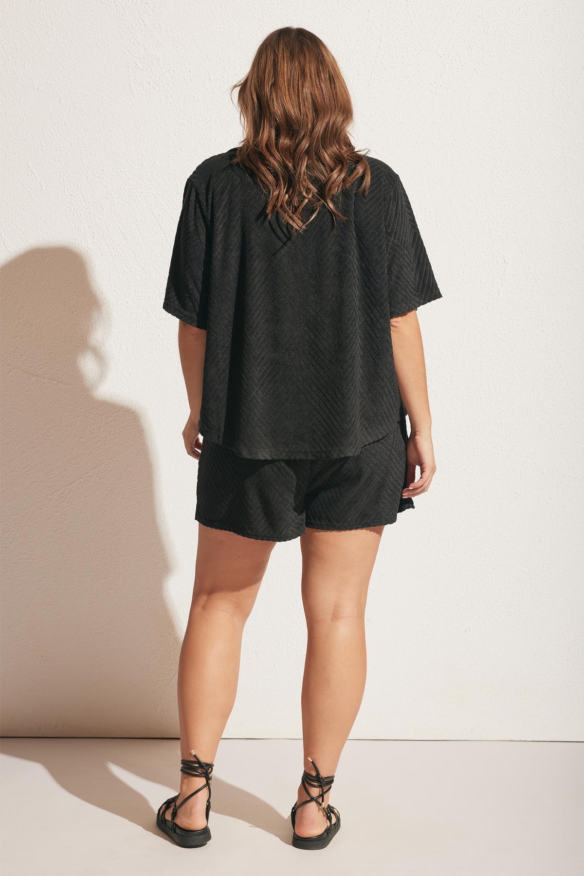 Woman seen from behind wearing a relaxed black terry shirt and matching shorts with lace-up sandals against a plain backdrop