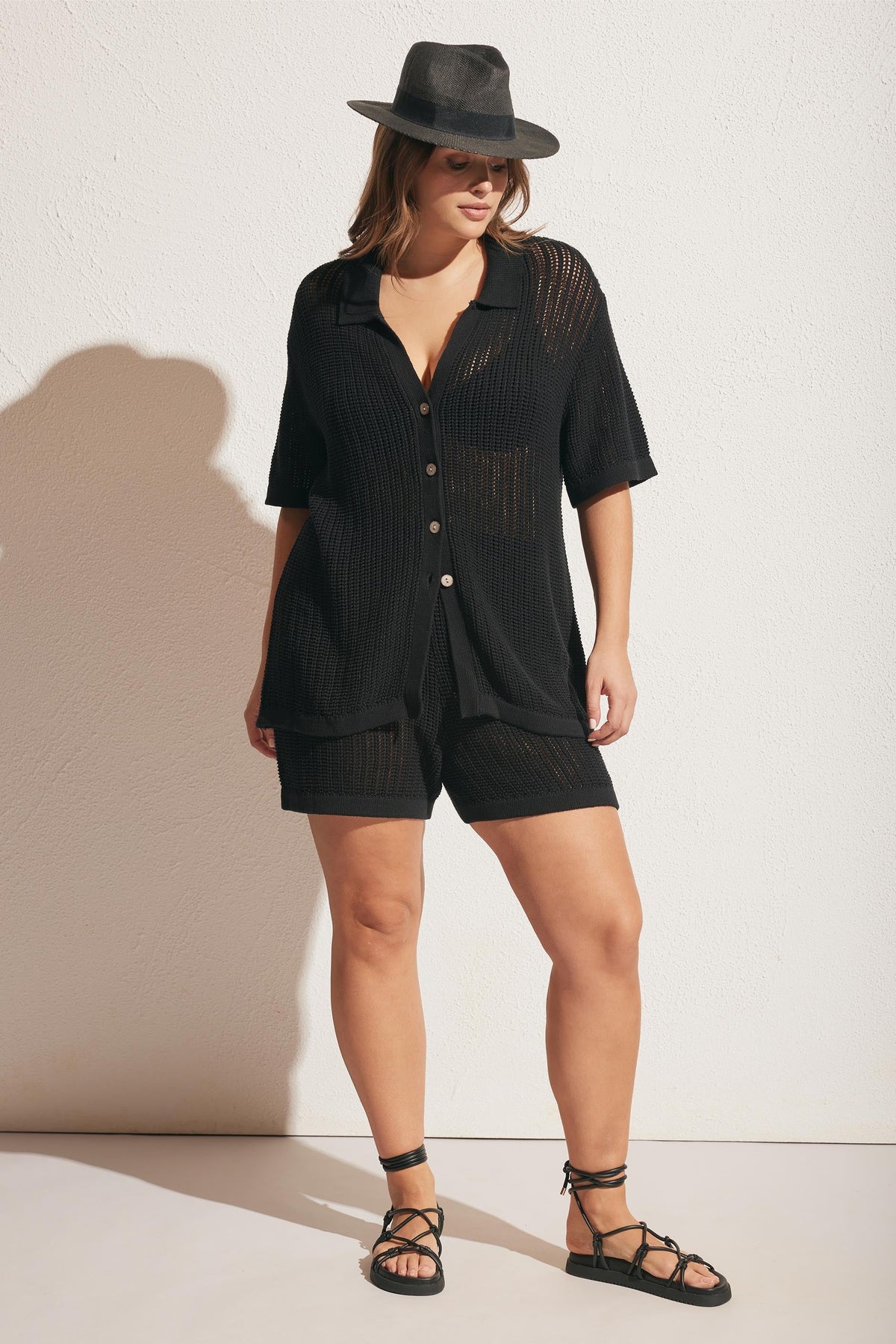 Woman wearing a black crochet button-up shirt with matching shorts, paired with a dark wide-brim fedora and strappy sandals