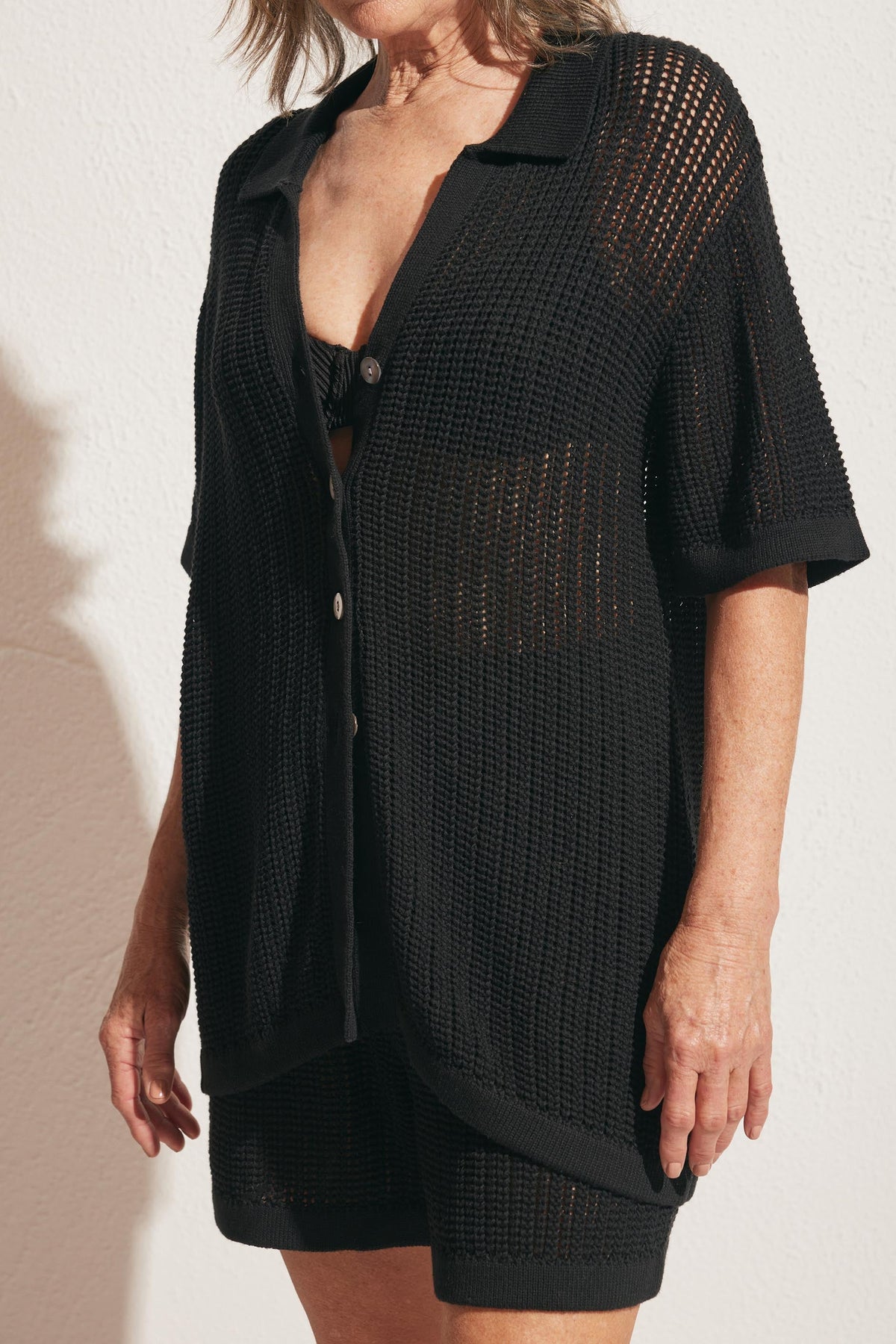 Woman wearing a black open-knit short-sleeve shirt over matching shorts and a black bandeau top against a light wall