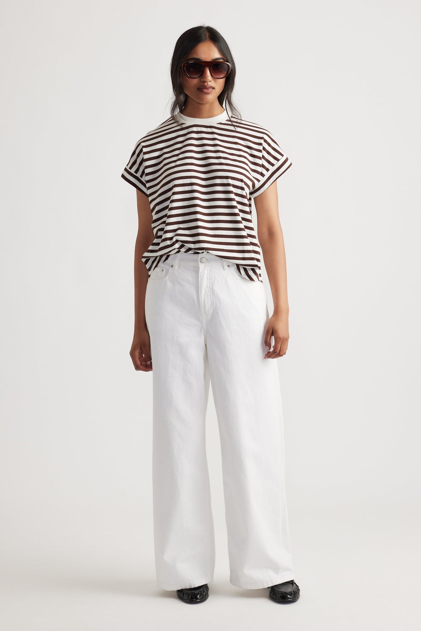 Woman wearing a brown and white striped tee shirt and white wide leg jeans on a plain background