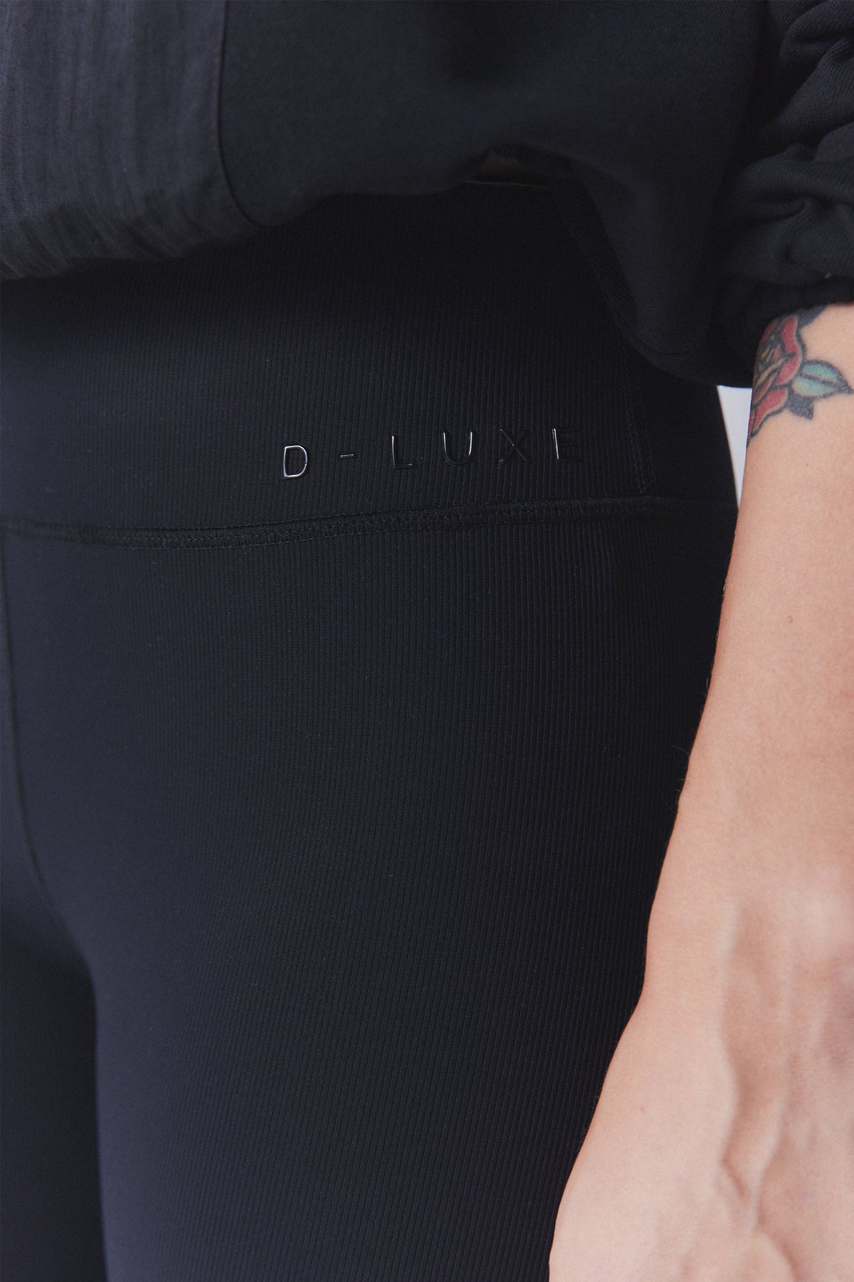 Close-up of black ribbed leggings waistband with subtle embossed branding and textured fabric detail on model's hip.