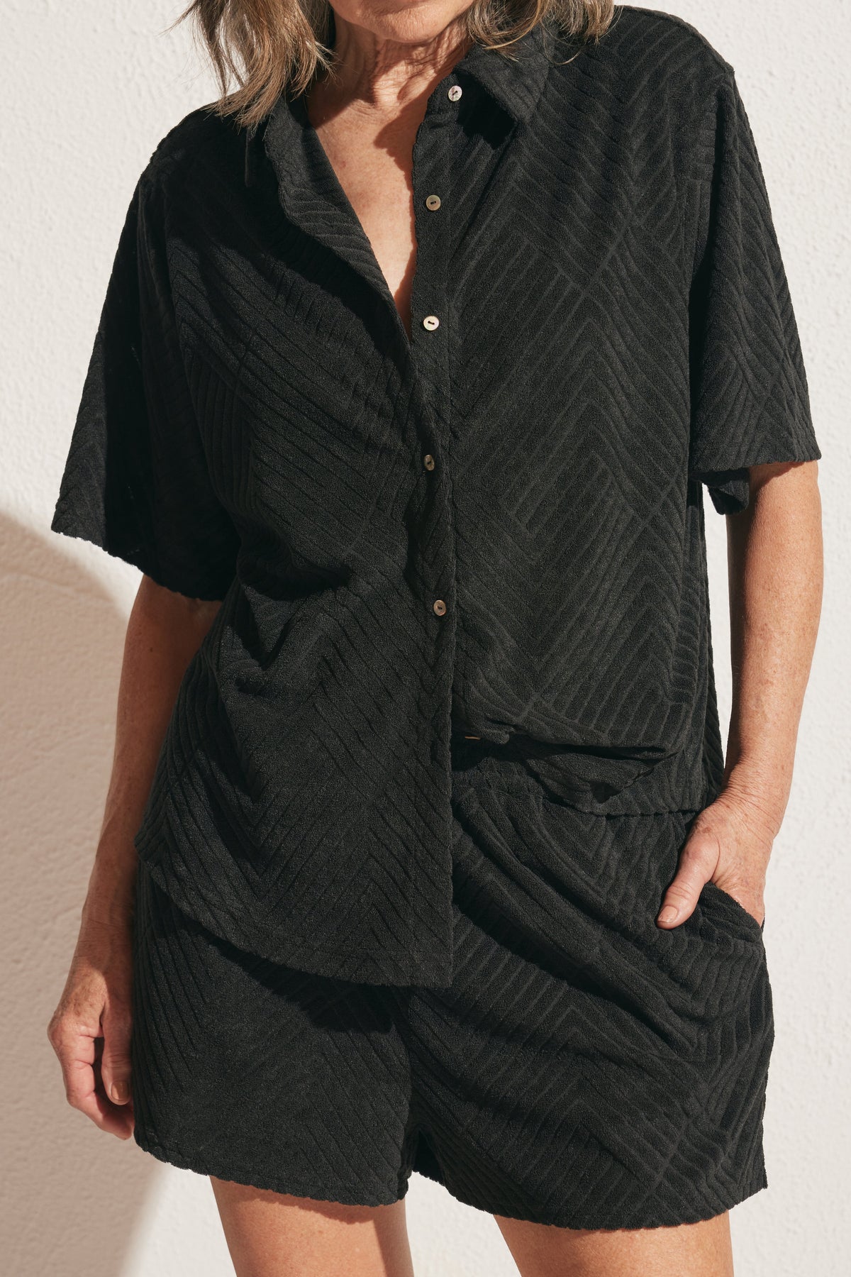Woman wearing a relaxed black terry shirt with subtle textured chevron pattern and matching shorts, hands in pockets