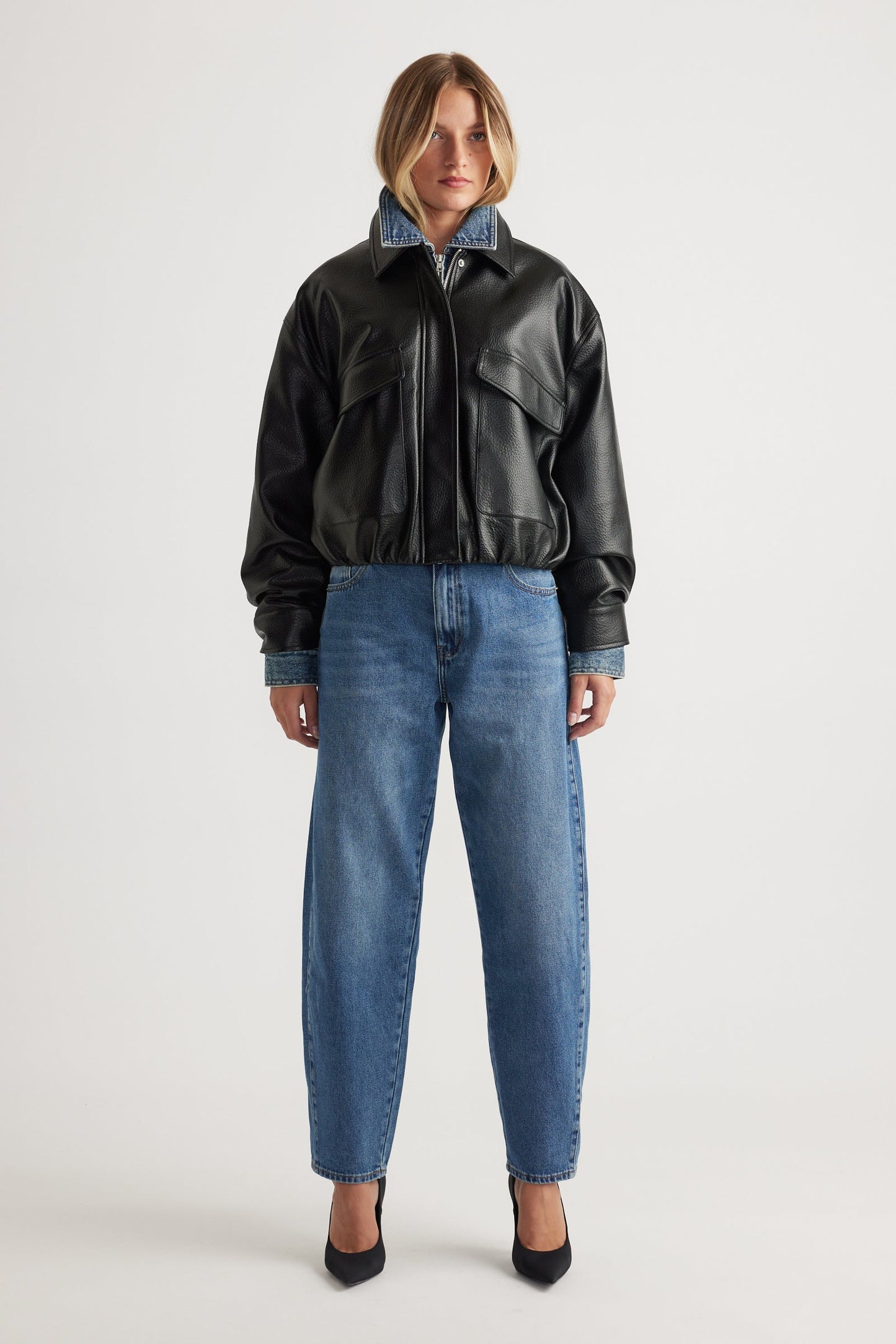 Woman wearing a black leather bomber jacket and mid wash blue barrel leg jeans on a white background
