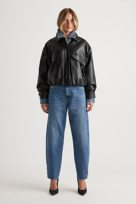 Woman wearing a black leather bomber jacket and mid wash blue barrel leg jeans on a white background
