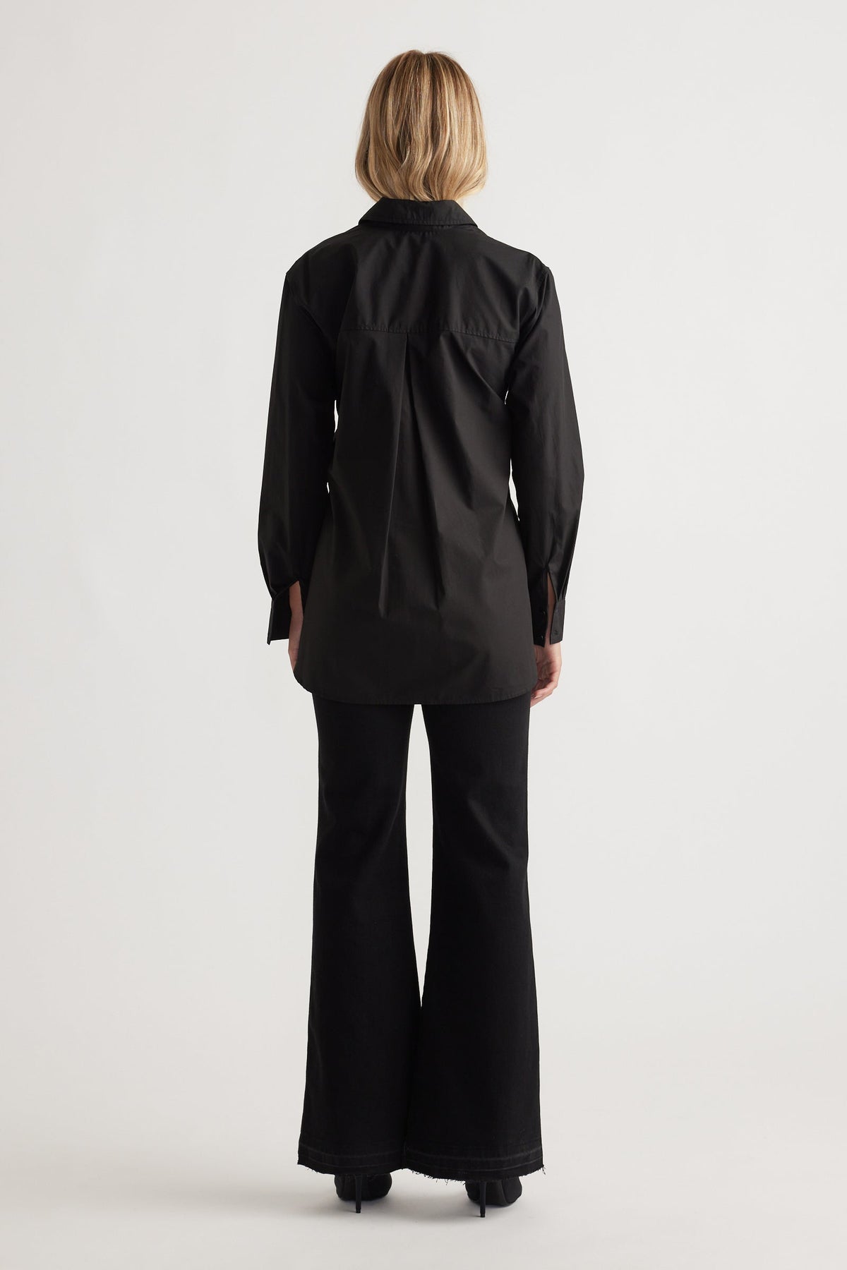 Back view of a woman wearing a sleek black tailored shirt with long sleeves paired with flared black pants and heels