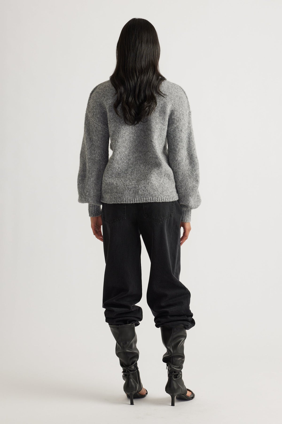 Woman seen from behind wearing a soft grey jumper with puff sleeves, black relaxed-fit pants, and slouchy heeled boots
