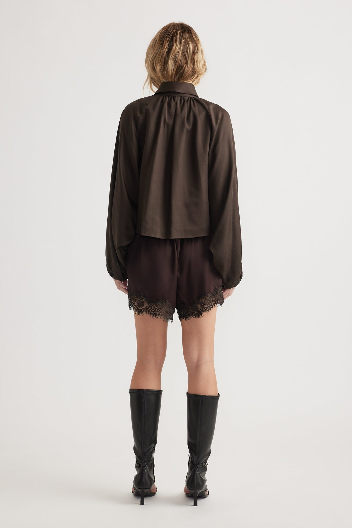 Woman wearing a loose dark long-sleeve top paired with black lace-trimmed shorts and knee-high boots, back view