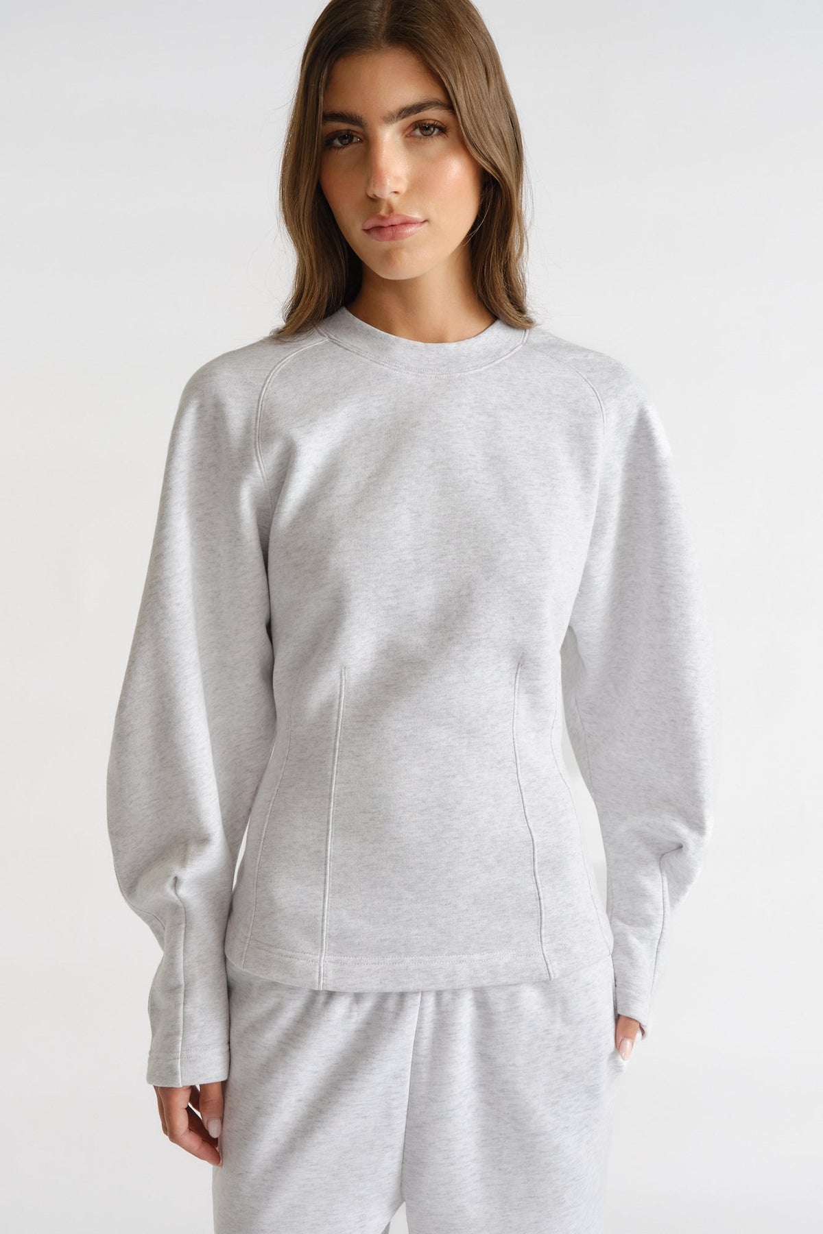 Woman wearing light grey cinched sweatshirt with long balloon sleeves and matching sweatpants against plain background