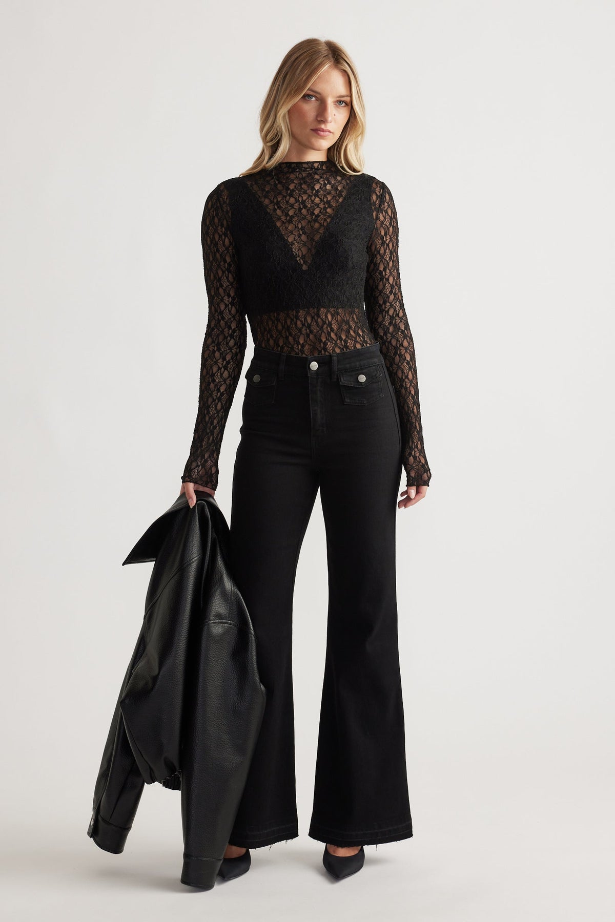 Woman wearing a black lace long sleeve top layered over a bralette, paired with high-waisted black flared jeans and holding a leather jacket