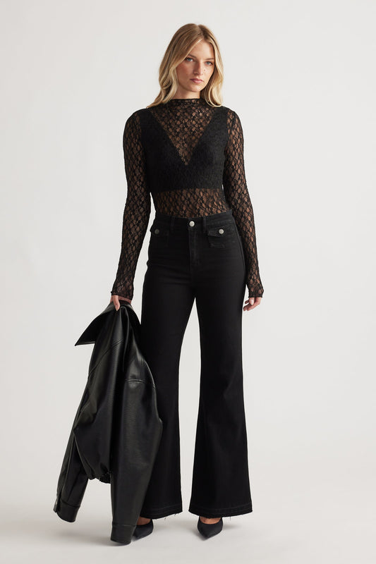 Woman wearing a black lace long sleeve top layered over a bralette, paired with high-waisted black flared jeans and holding a leather jacket