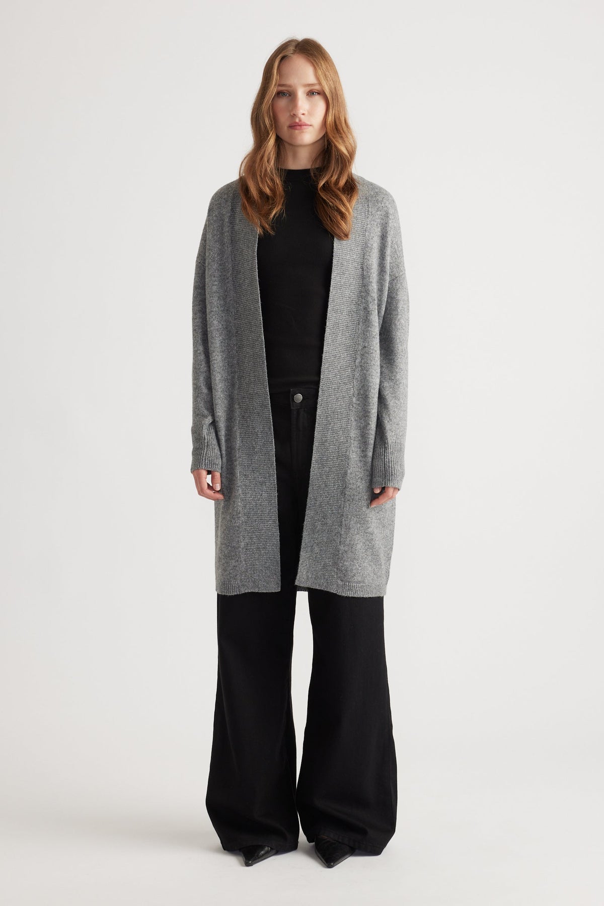 Woman wearing mid-grey longline knit cardigan over black top and wide-leg black pants against white background