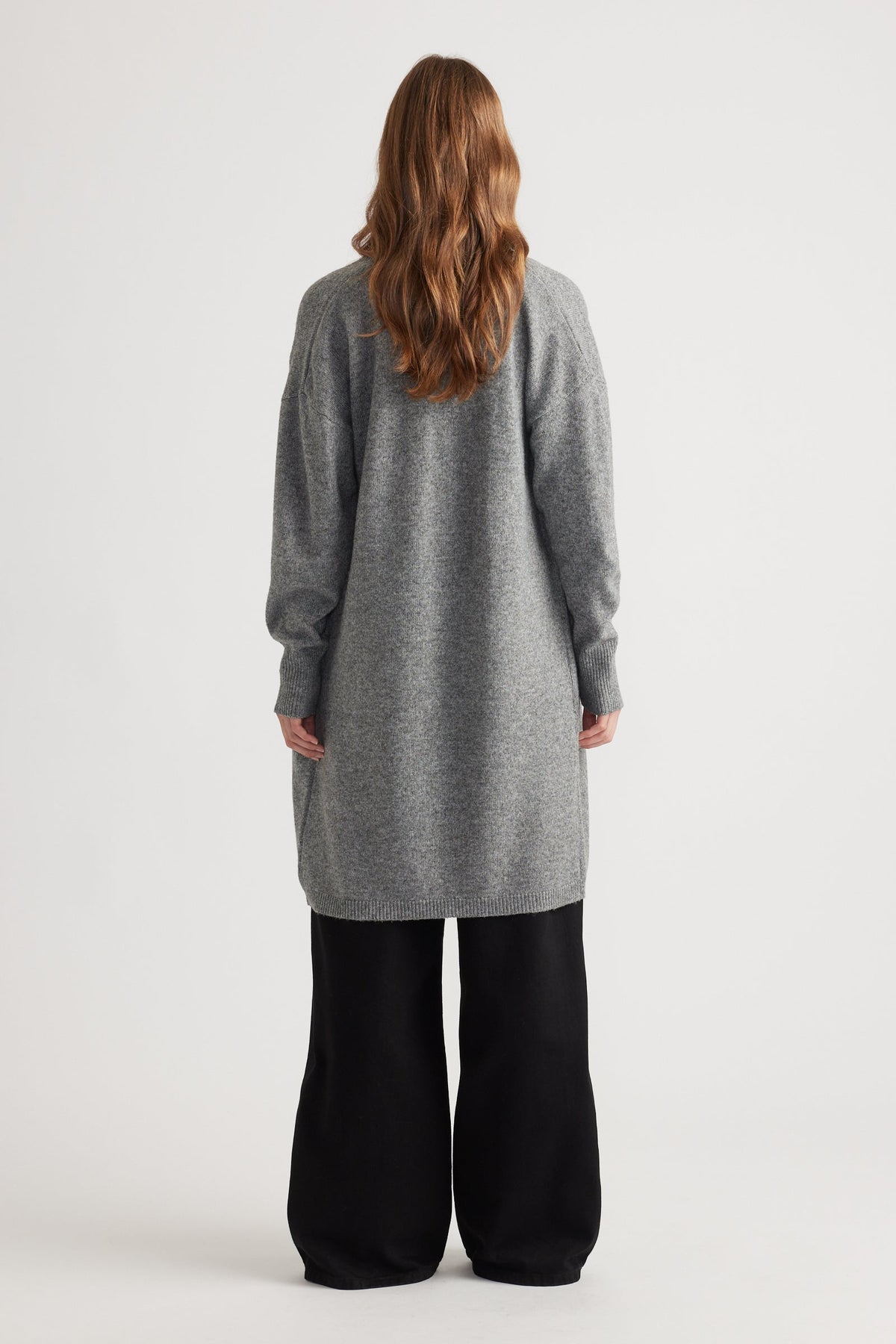 Woman shown from behind wearing a mid-grey longline knit cardigan paired with loose black pants against a plain backdrop