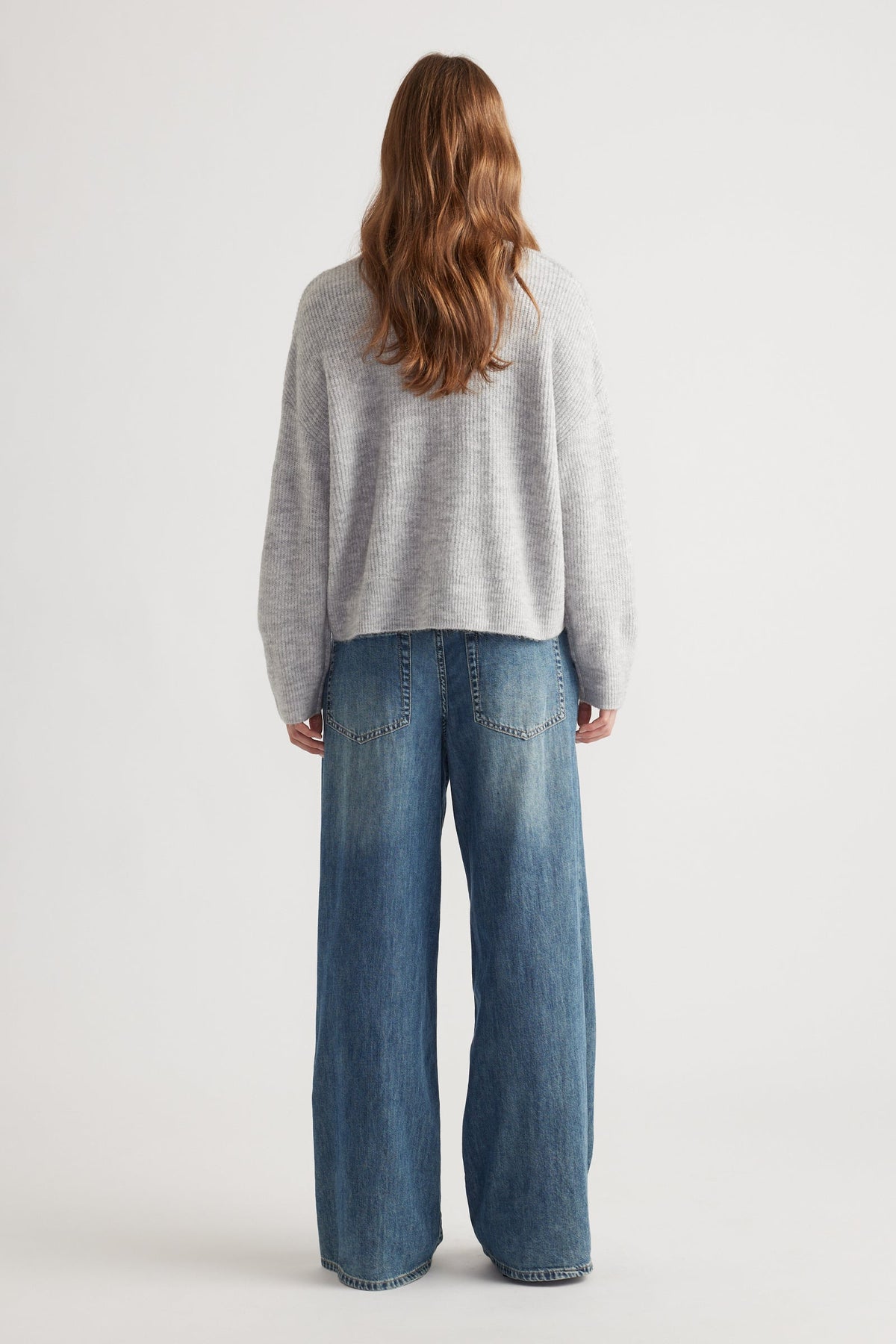 Woman with loose waves wearing a cozy light grey knit cardigan paired with wide-leg faded blue denim jeans viewed from behind