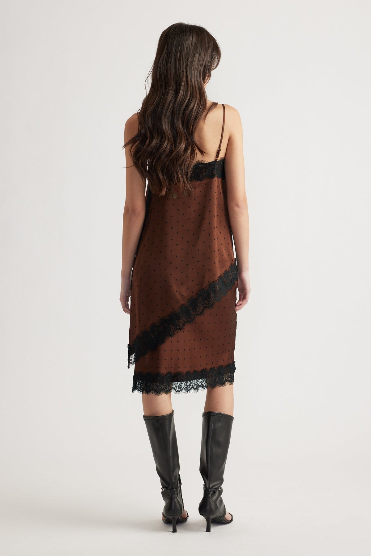 Woman wearing a brown slip dress with black lace trim, thin straps, and knee-high black heeled boots viewed from behind