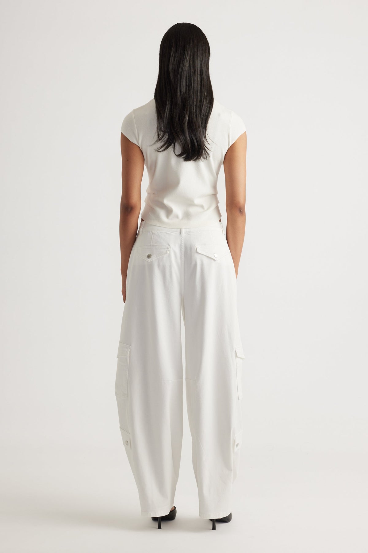 Rear view of a woman wearing the Lily Pleat Detail Knit Tee in soft winter white paired with wide-leg white pants by DECJUBA