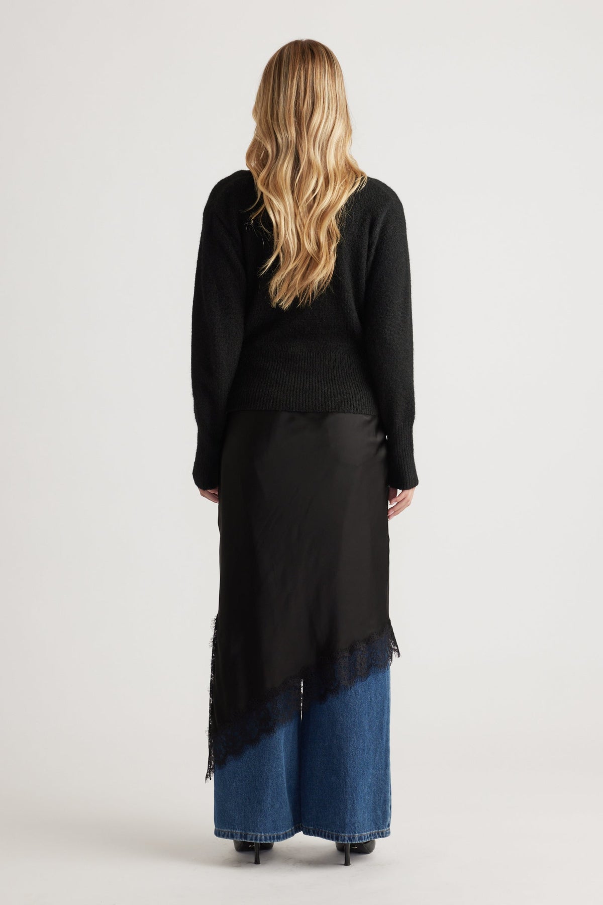 Back view of a black knit crossover cardigan layered over a silky asymmetrical lace-trimmed skirt paired with wide-leg blue denim jeans