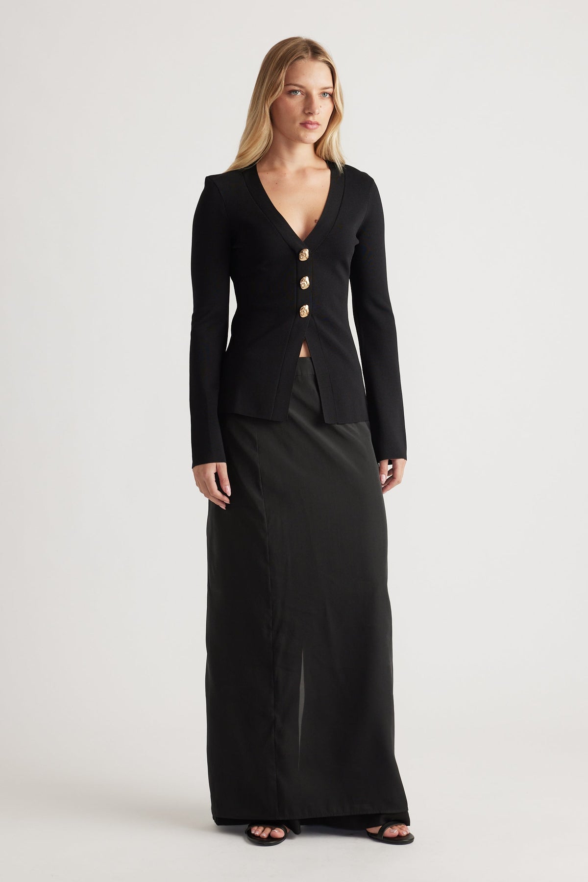Woman wearing a fitted black knit cardigan with gold buttons and deep V-neck, paired with a long black skirt