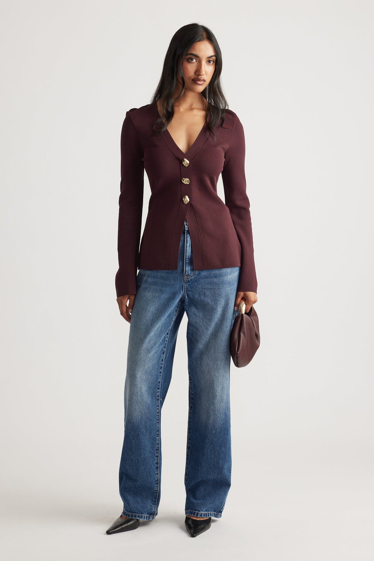 Woman wearing a fitted burgundy knit cardigan with gold buttons, paired with relaxed blue denim jeans and black pointed shoes.