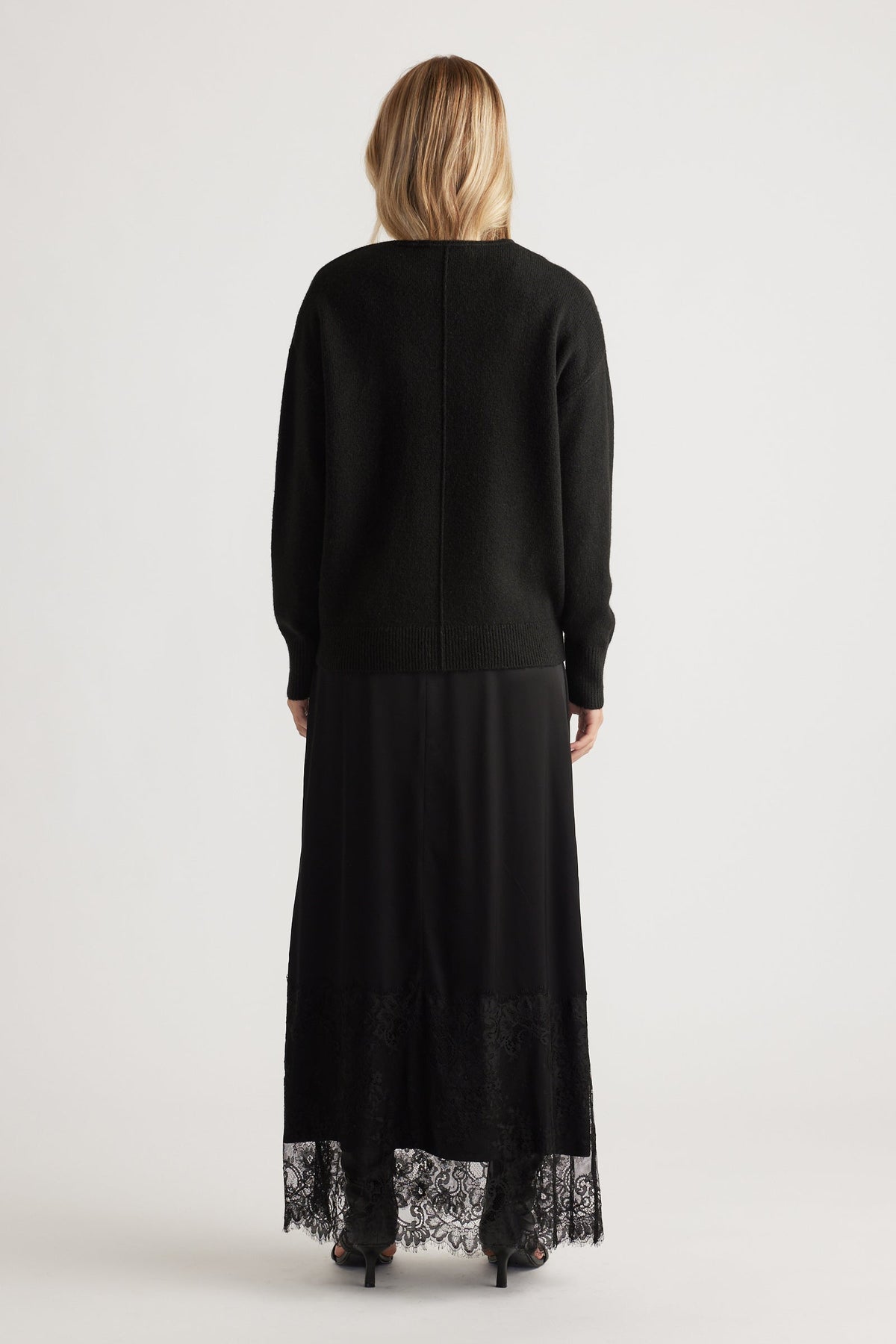 Back view of woman wearing a black knit jumper with seam detail paired with a long black lace-trimmed skirt and heels