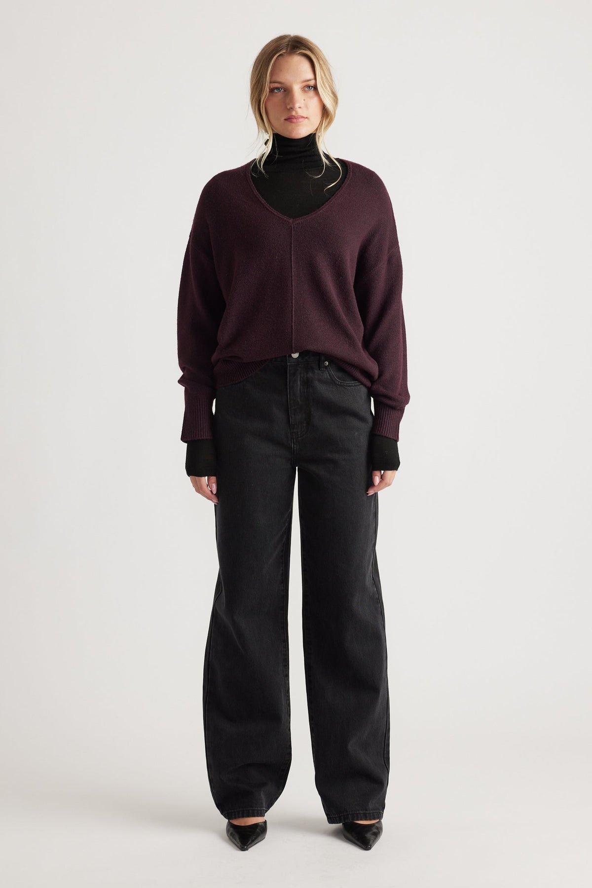 Woman wearing a relaxed-fit burgundy V-neck knit jumper with central seam over a black turtleneck and wide-leg black jeans