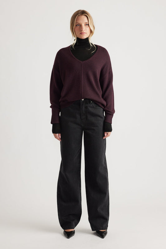 Woman wearing a relaxed-fit burgundy V-neck knit jumper with central seam over a black turtleneck and wide-leg black jeans