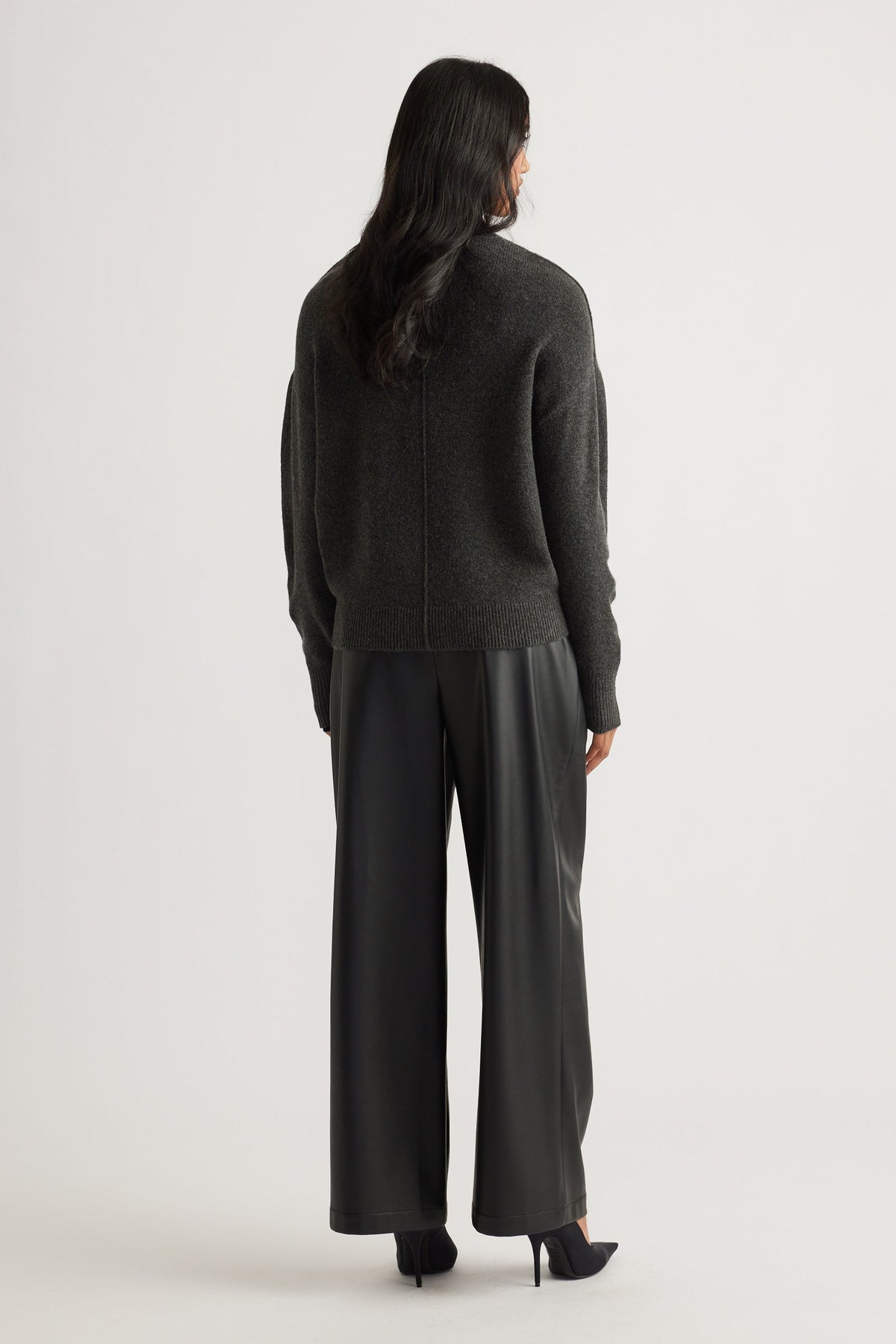 Woman wearing a charcoal knit jumper with subtle seam detail, paired with black wide-leg leather-look pants and heels