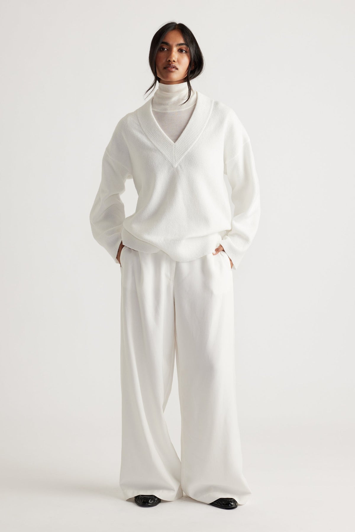 Model dressed in a soft white V-neck knit jumper layered over a high-neck top, paired with wide-leg trousers.