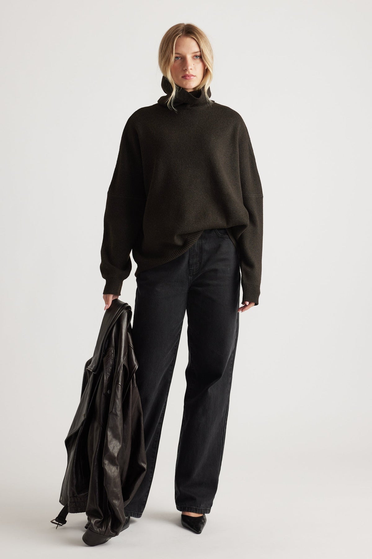 Woman wearing a dark roll neck knit jumper paired with relaxed black jeans and holding a leather jacket for a casual look