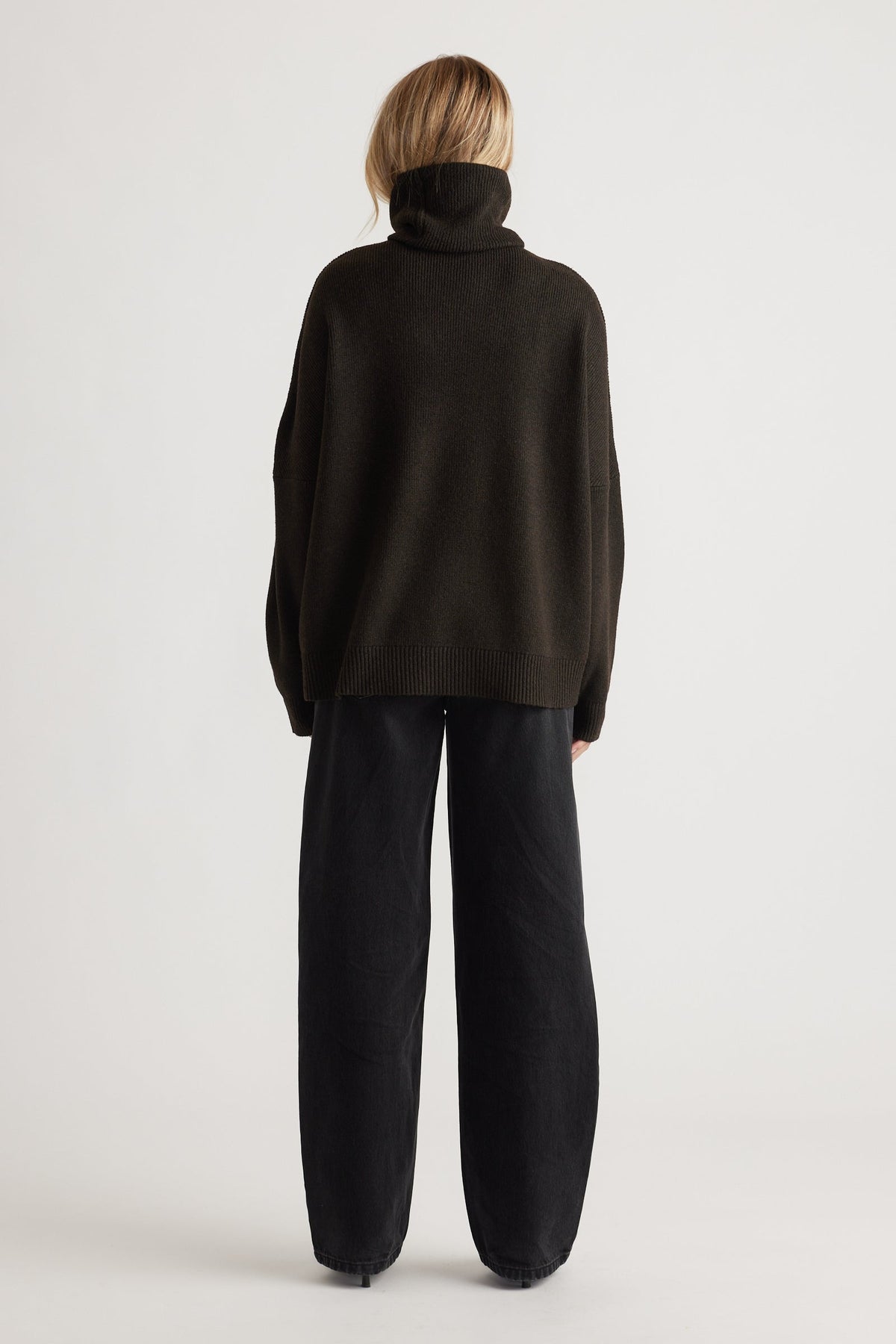 Woman wearing a loose dark brown roll neck jumper paired with relaxed black wide-leg pants in a minimalist setting