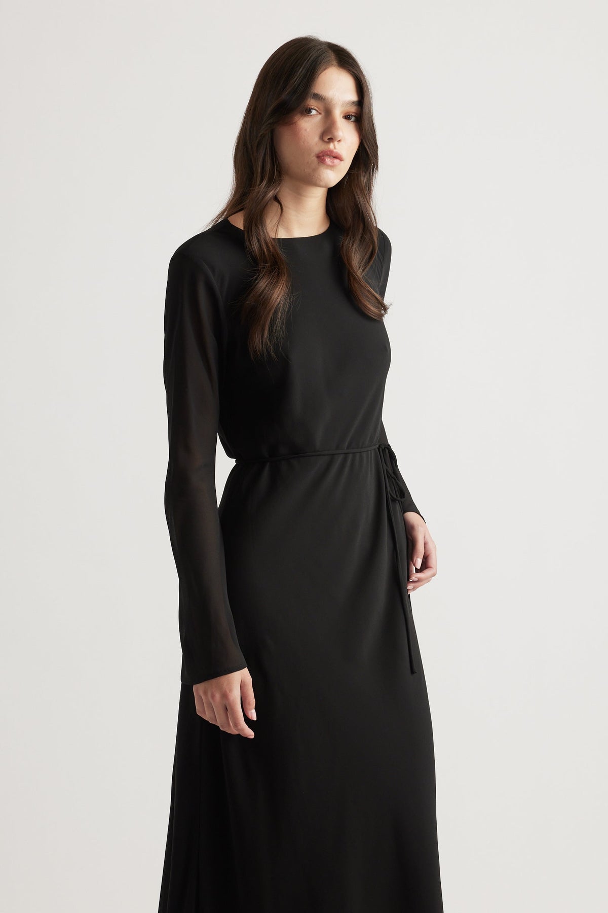Woman wearing a sleek black maxi dress with sheer long sleeves and a waist tie for a refined, modern look