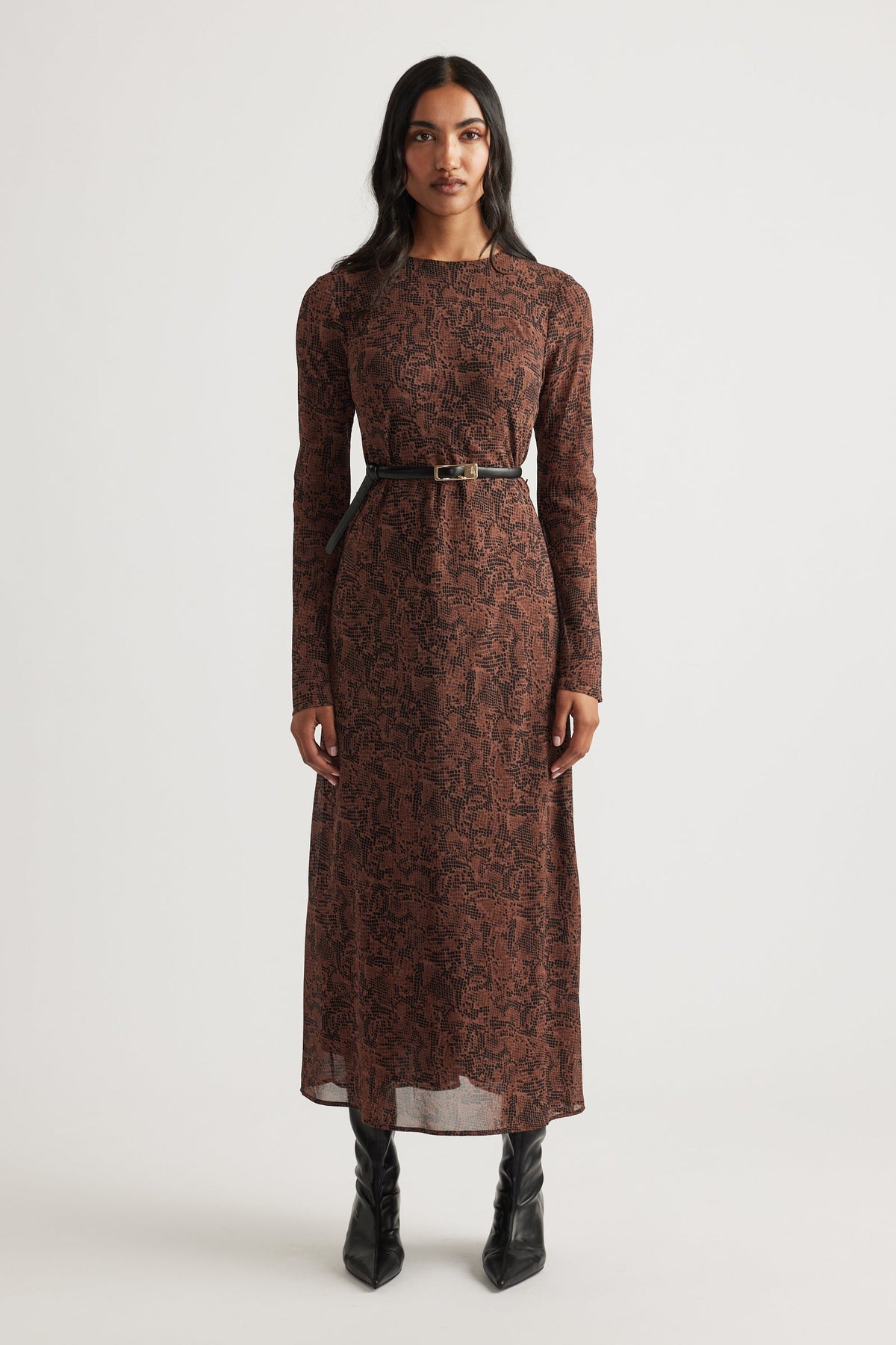 Woman wearing a long-sleeve maxi dress with a brown abstract pattern, cinched at the waist with a slim black belt and black boots