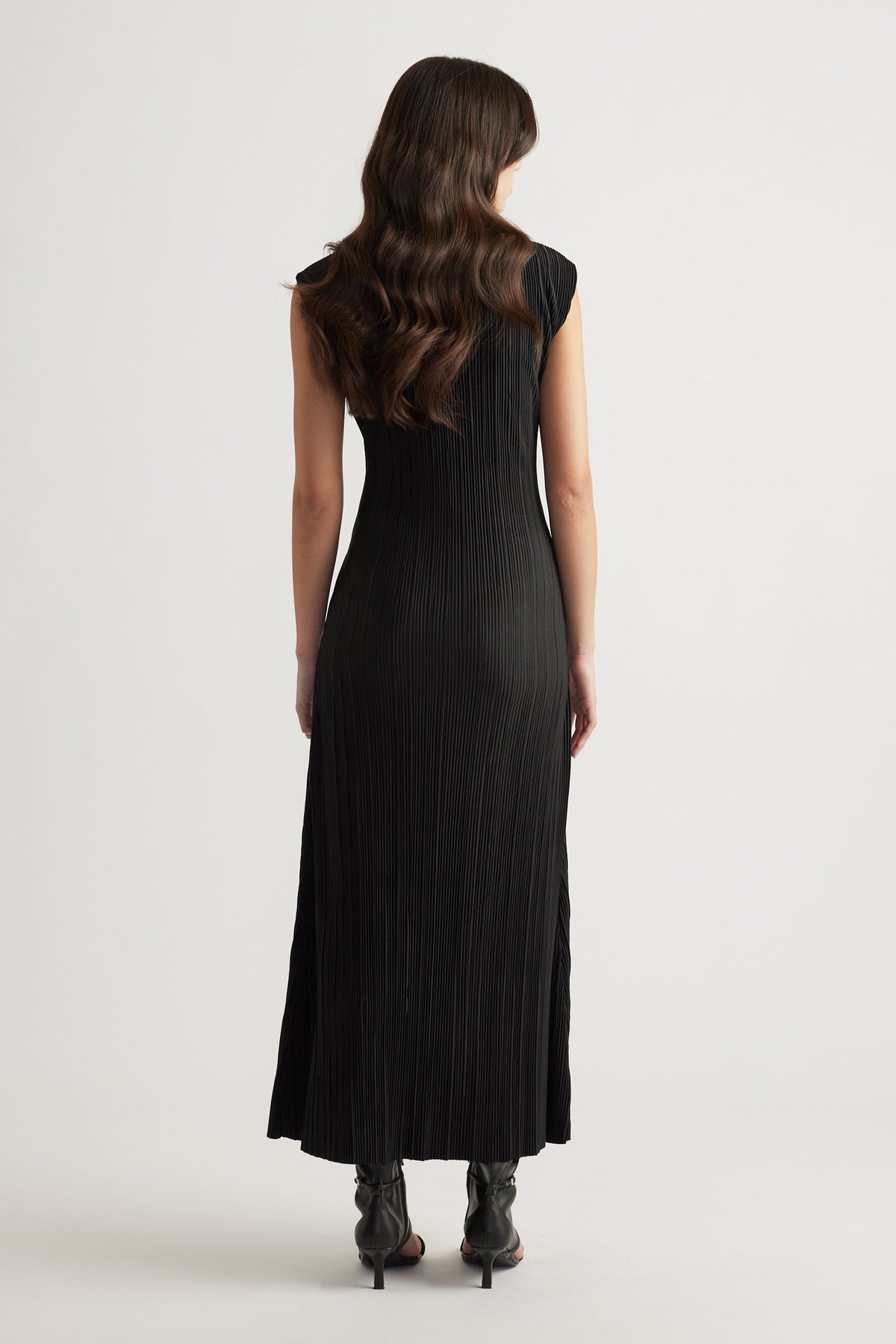 Woman wearing a sleek black pleated maxi dress with cap sleeves and a flowing silhouette paired with black heeled boots.
