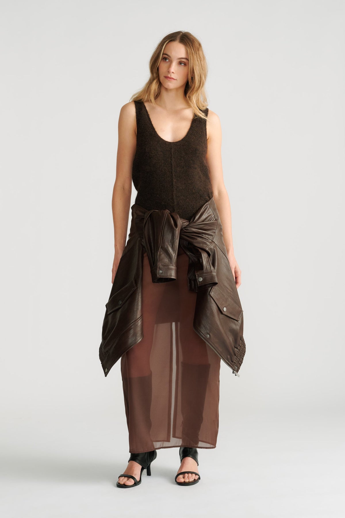 Woman wearing a dark brown fluffy knit tank with a tied leather jacket and sheer brown maxi skirt paired with black heels