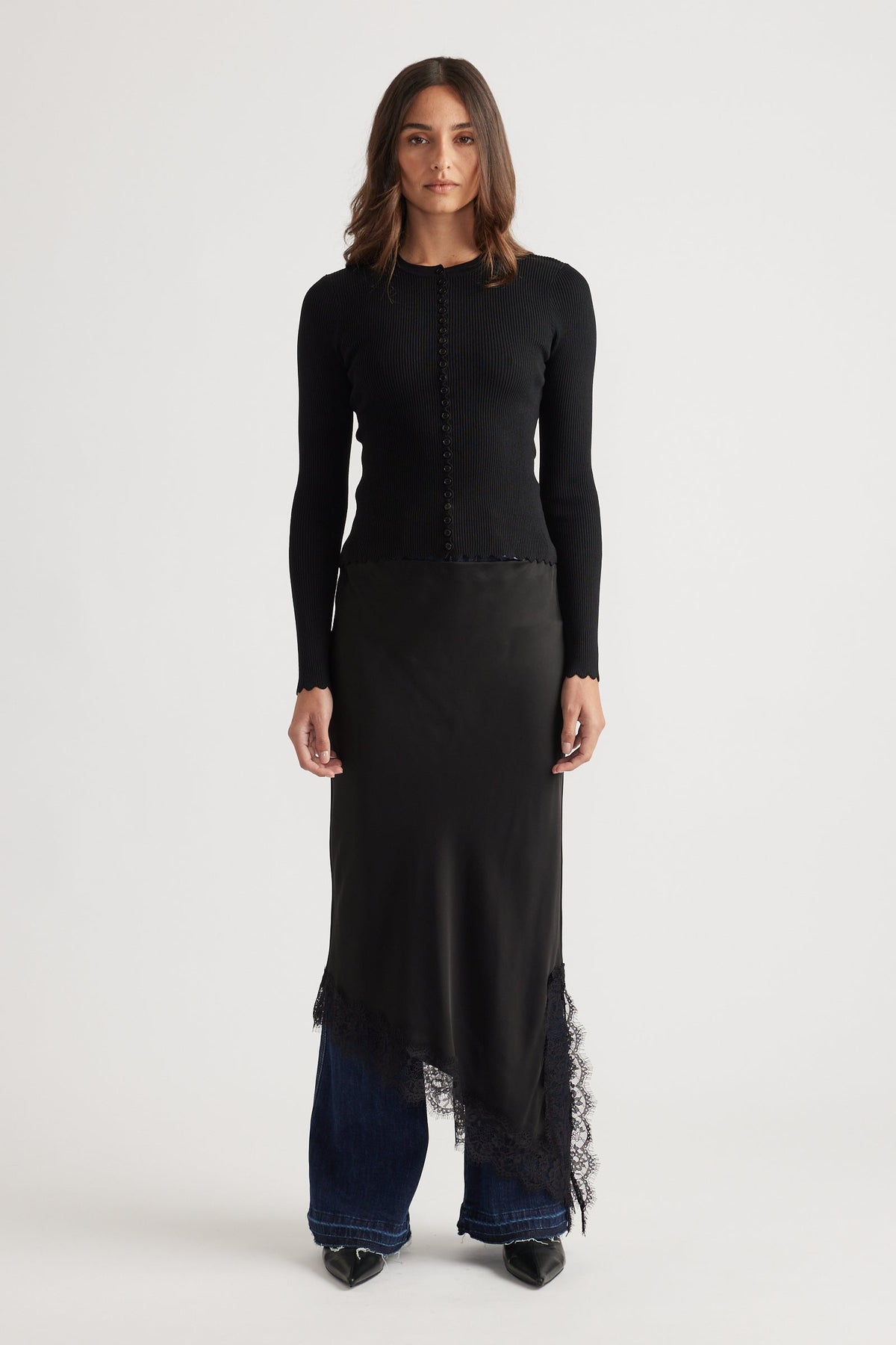 Woman wearing a fitted black top with scalloped button details paired with an asymmetrical lace-trimmed skirt and jeans