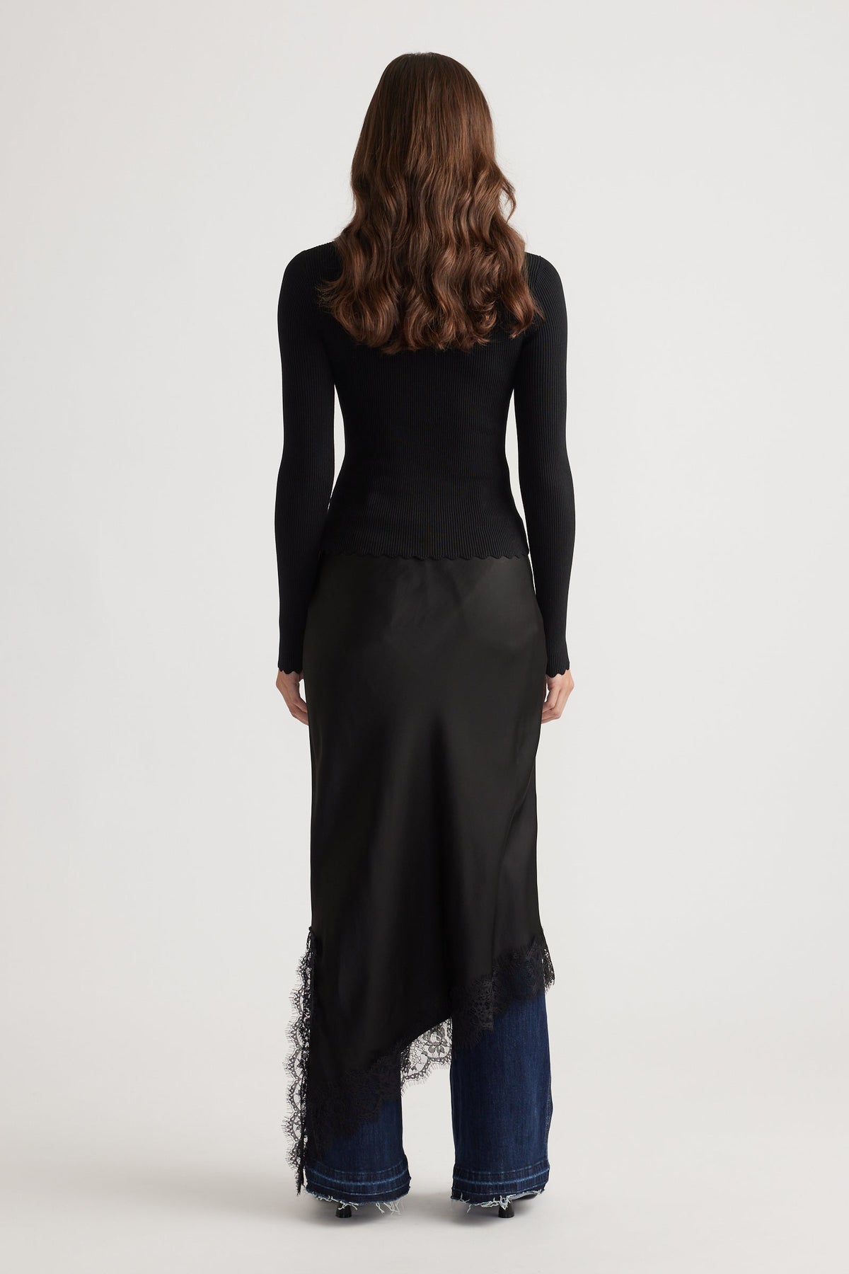 Slim-fit black top with scalloped hem and long sleeves paired with a lace-trimmed asymmetrical skirt layered over wide-leg denim jeans