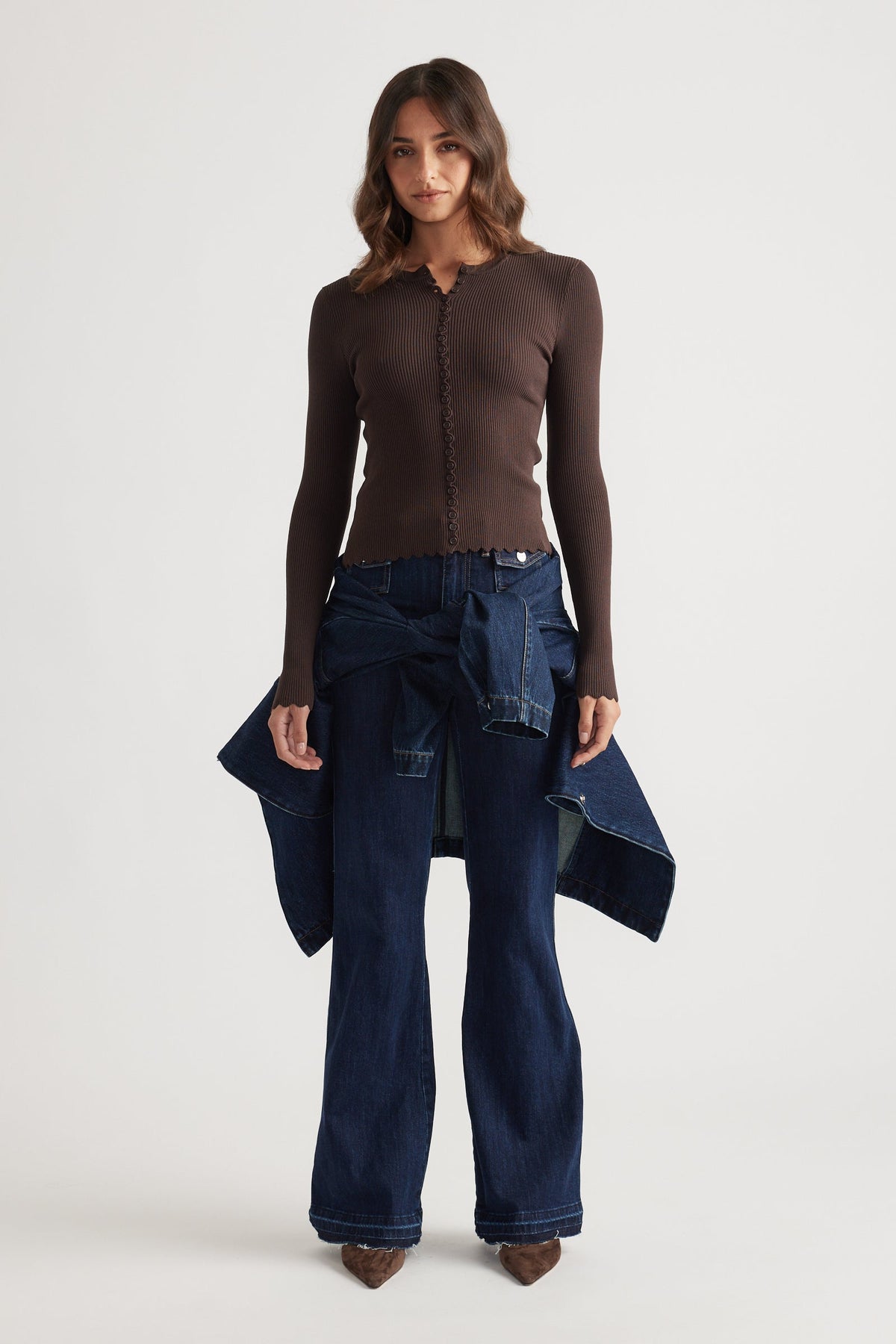 Woman wearing a fitted chocolate brown scalloped button long sleeve top with dark wide-leg denim jeans and brown pointed shoes