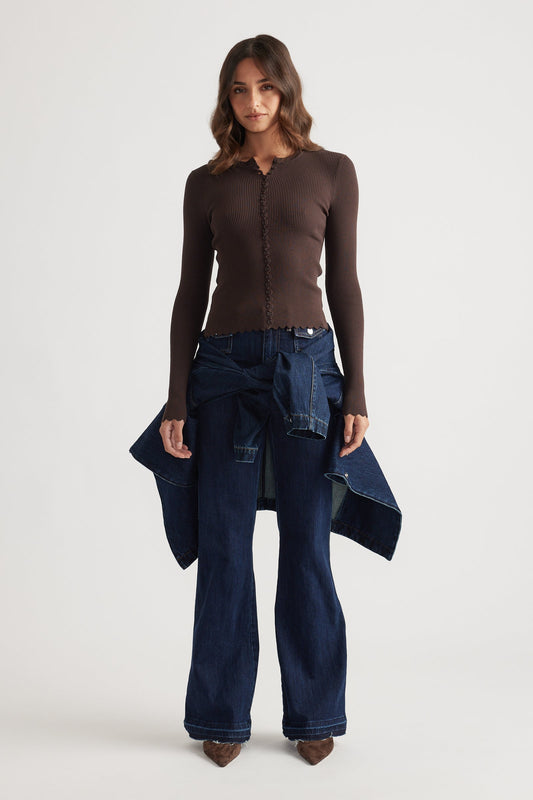 Woman wearing a fitted chocolate brown scalloped button long sleeve top with dark wide-leg denim jeans and brown pointed shoes