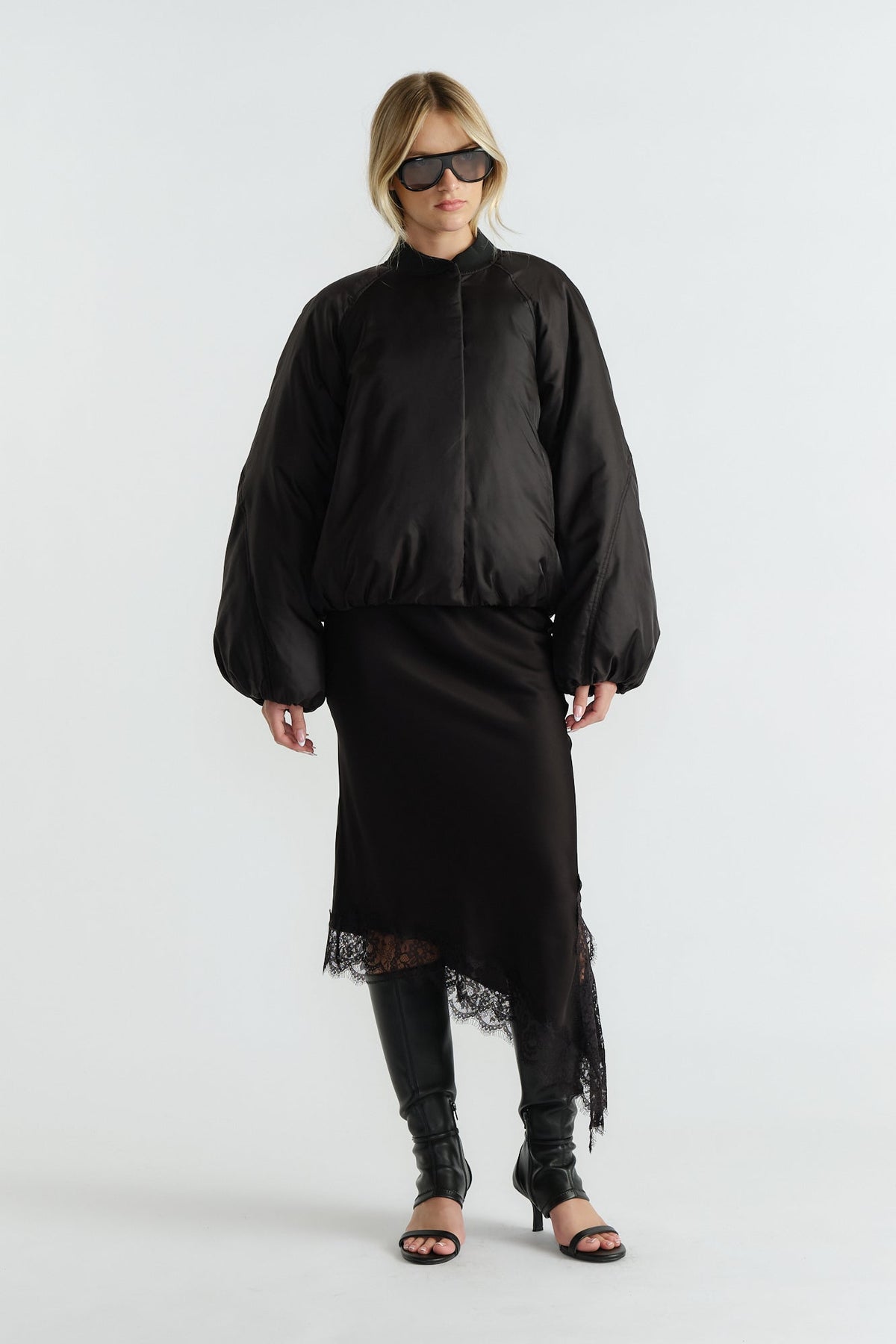 Woman wearing an oversized black bomber jacket with balloon sleeves over a black lace-trim midi skirt and heeled boots