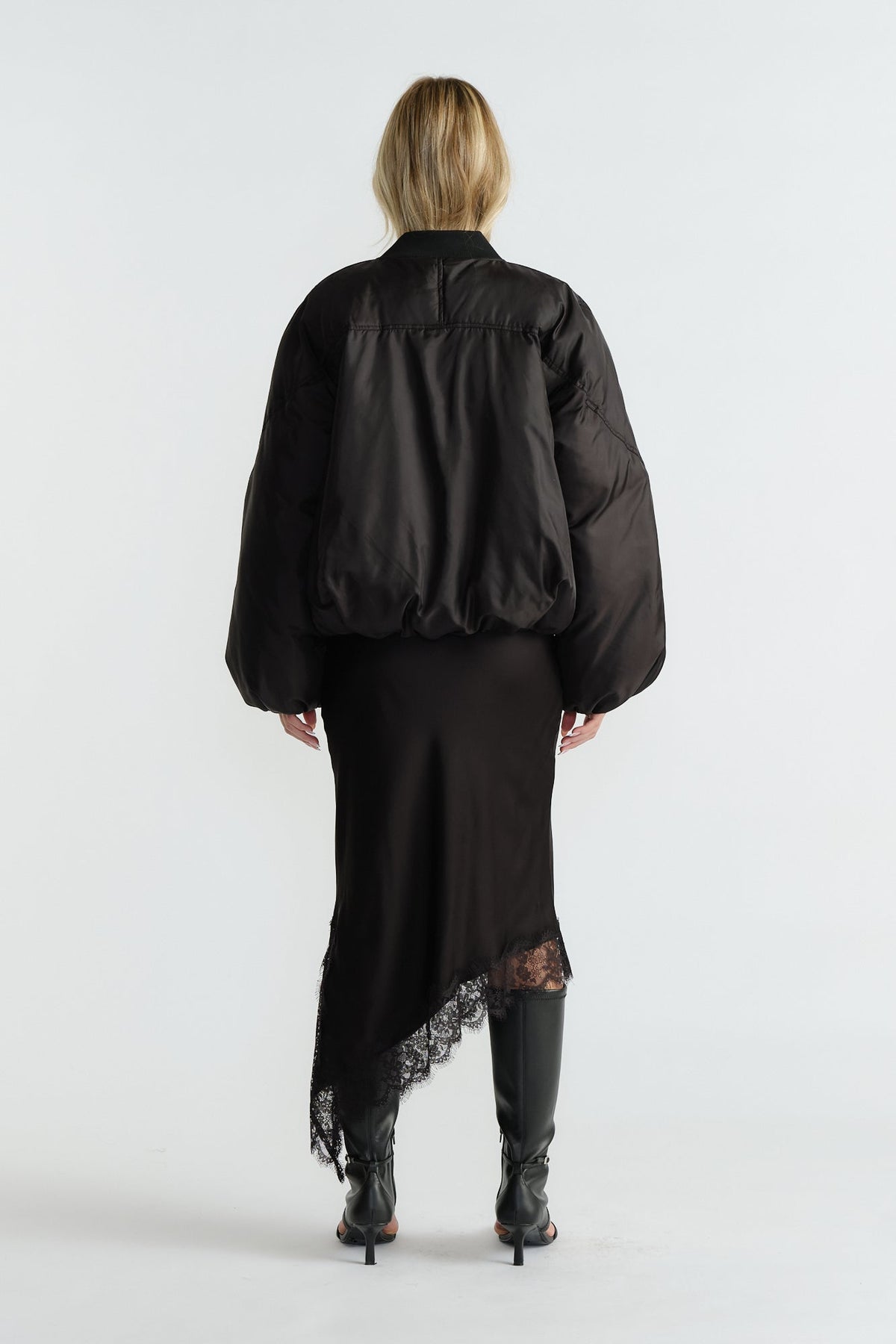Back view of woman wearing a black oversized bomber jacket paired with an asymmetric lace-hem skirt and black heeled boots