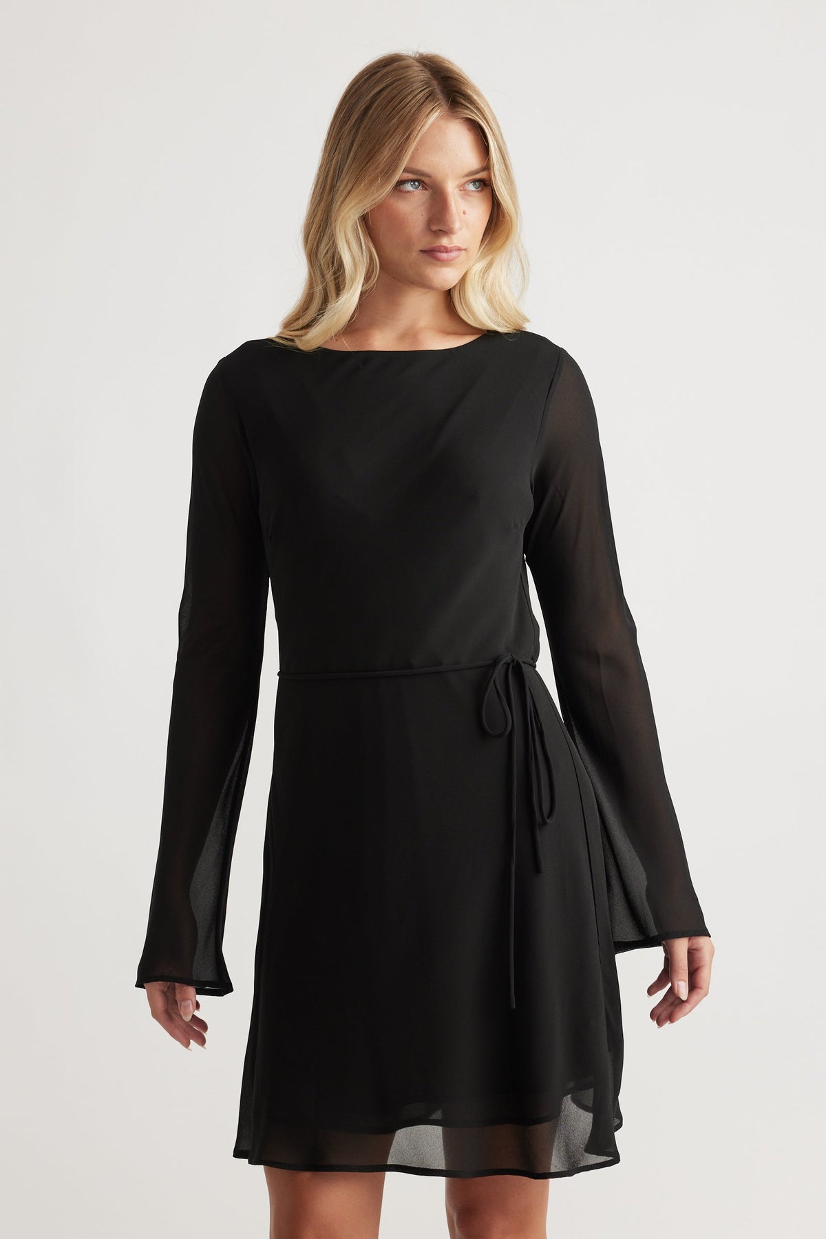 Woman wearing Chelsea Georgette Mini Dress in black with sheer long sleeves and a waist tie for a graceful look