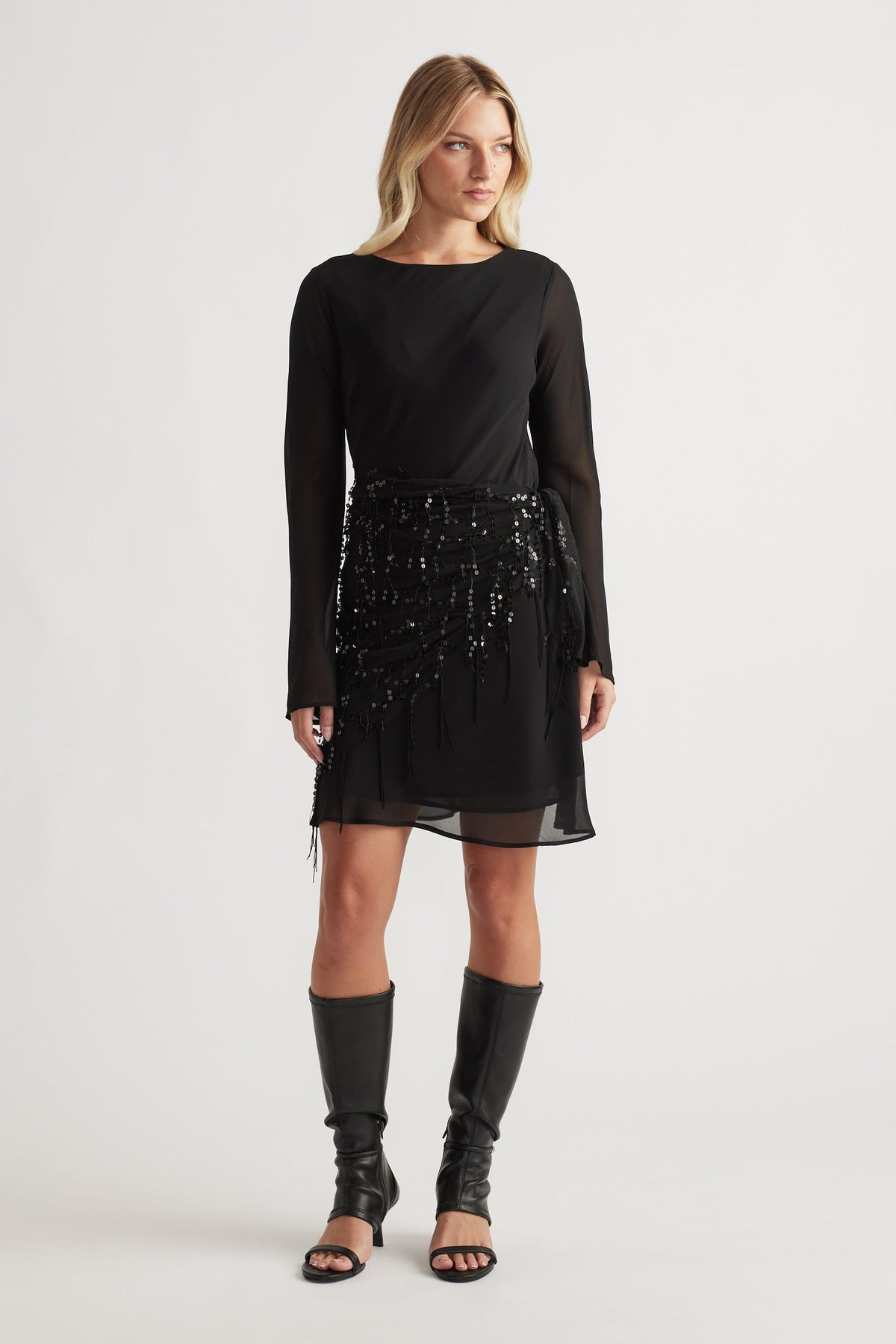 Model wearing Chelsea Georgette Mini Dress in black with sequin details, sheer long sleeves, paired with knee-high black boots