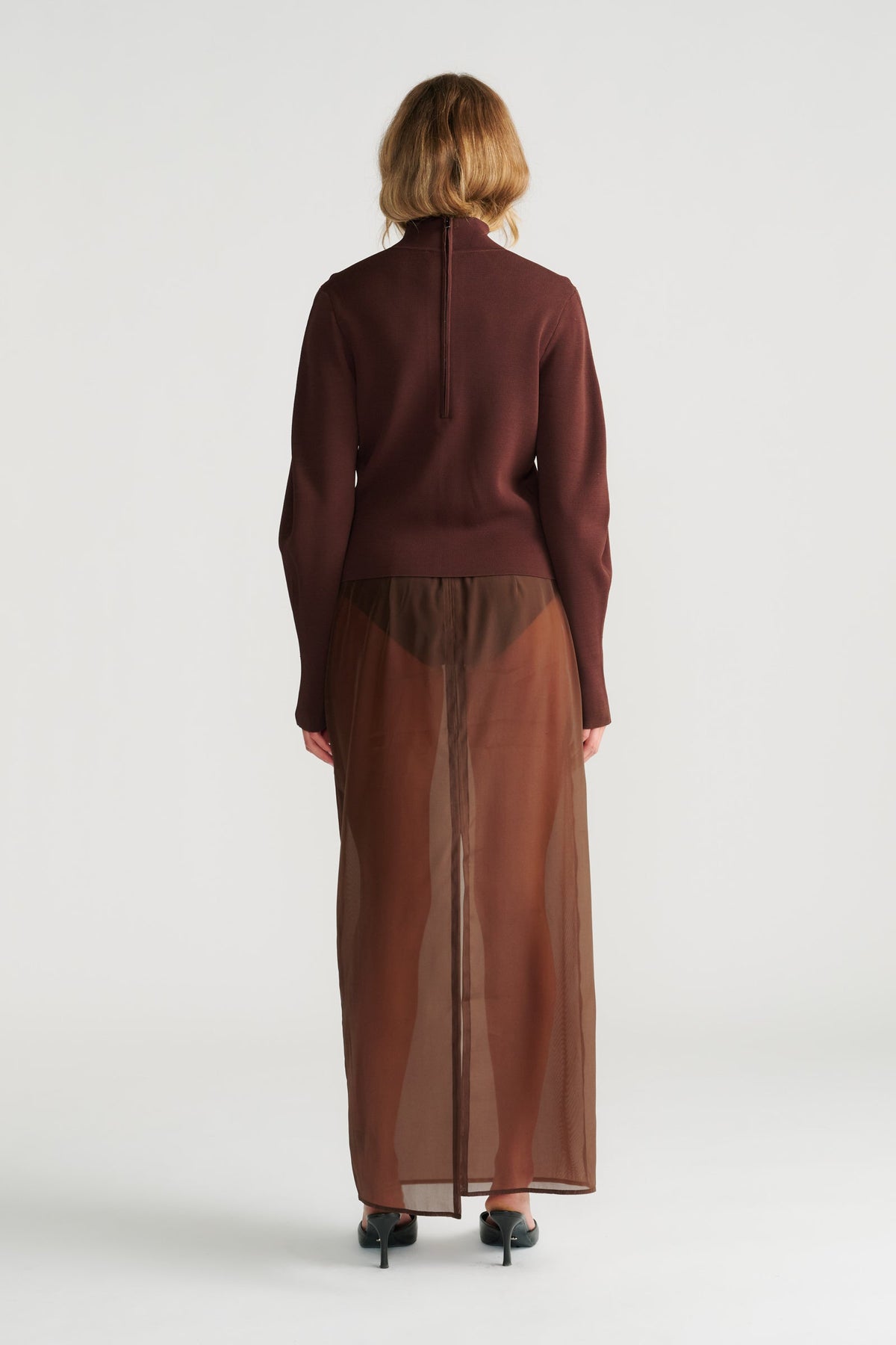 Woman wearing a rich mocha high neck jumper paired with a sheer brown maxi skirt and black heeled sandals, back view.